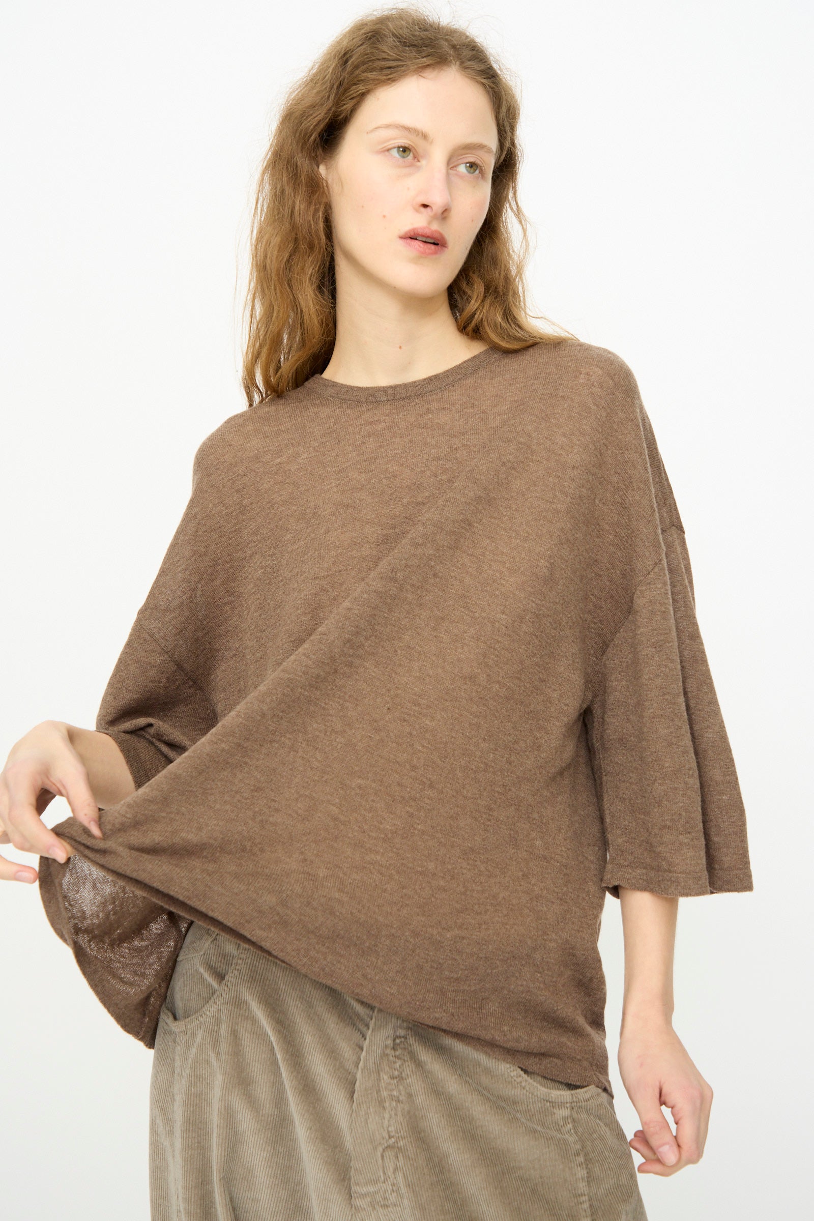 A person stands against a plain light background wearing Cordera’s Merino Wool Oversized T-Shirt in Deep Taupe, holding one side to show off its relaxed silhouette.