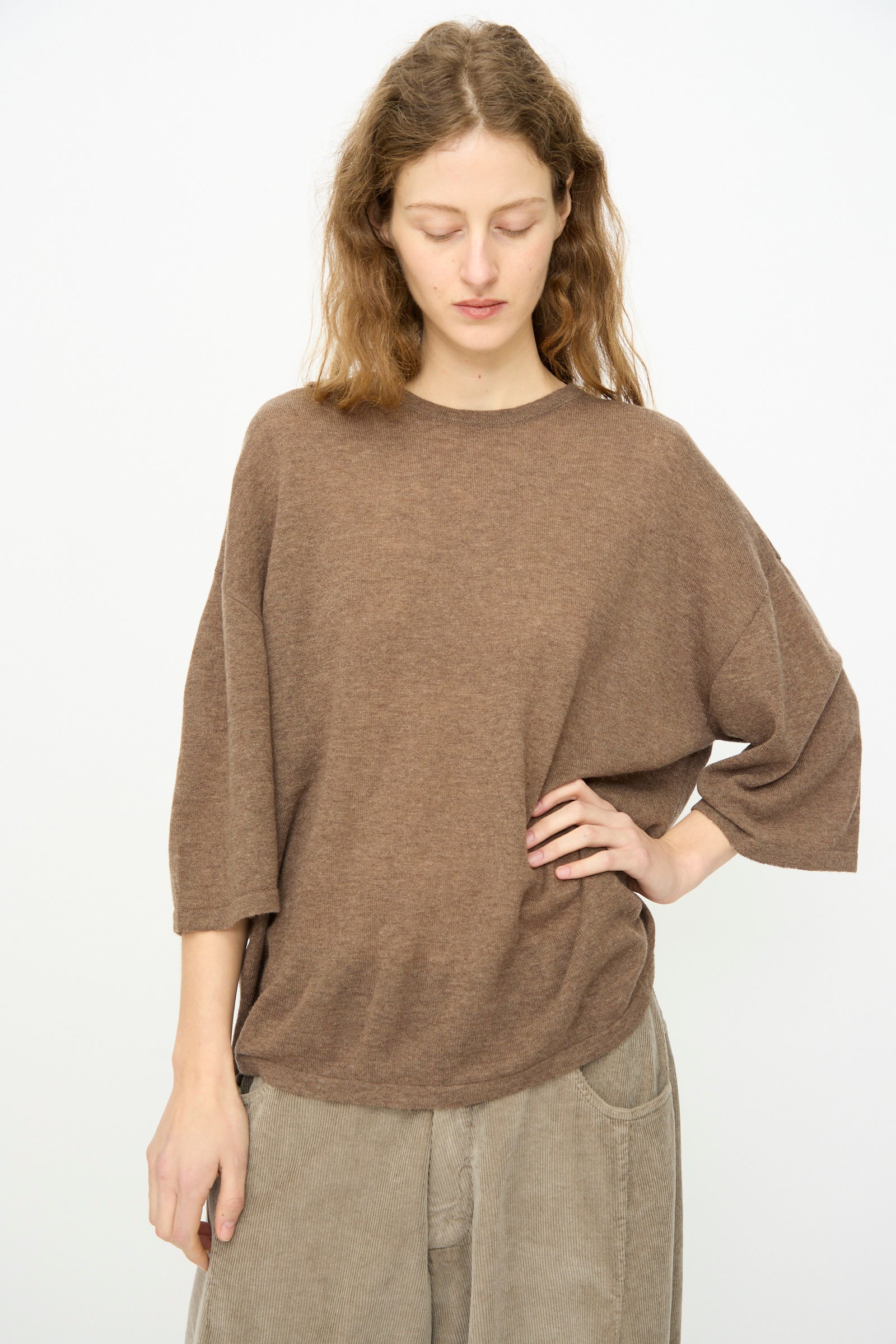 A person with wavy brown hair wears a relaxed silhouette: Cordera's Merino Wool Oversized T-Shirt in Deep Taupe and beige corduroy pants, standing against a plain white background and looking down with one hand on their hip.