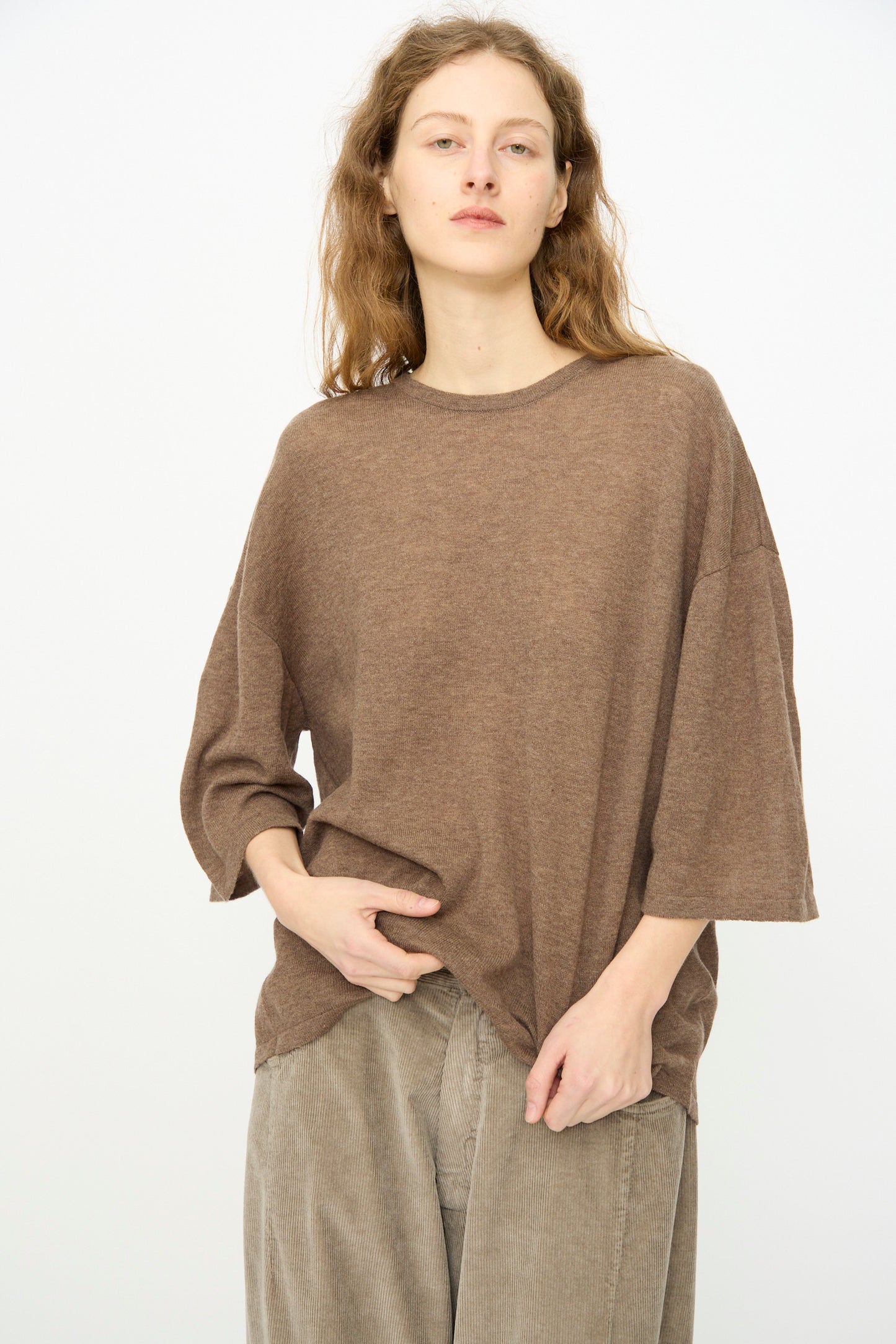 A person with wavy, light brown hair wears the Cordera Merino Wool Oversized T-Shirt in Deep Taupe and beige pants, standing against a plain white background.