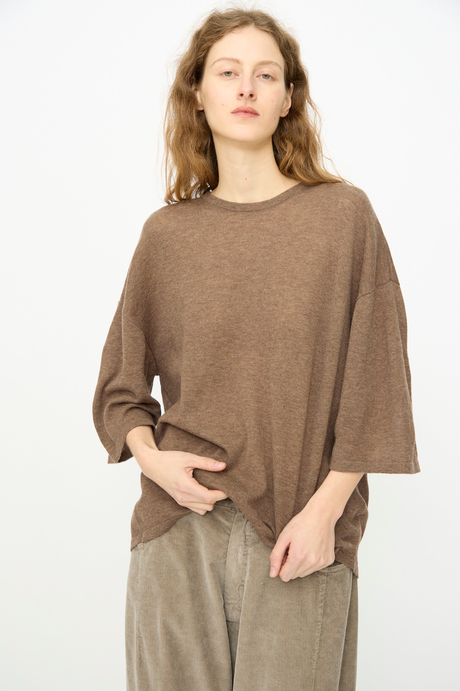 A person with wavy, light brown hair wears the Cordera Merino Wool Oversized T-Shirt in Deep Taupe and beige pants, standing against a plain white background.