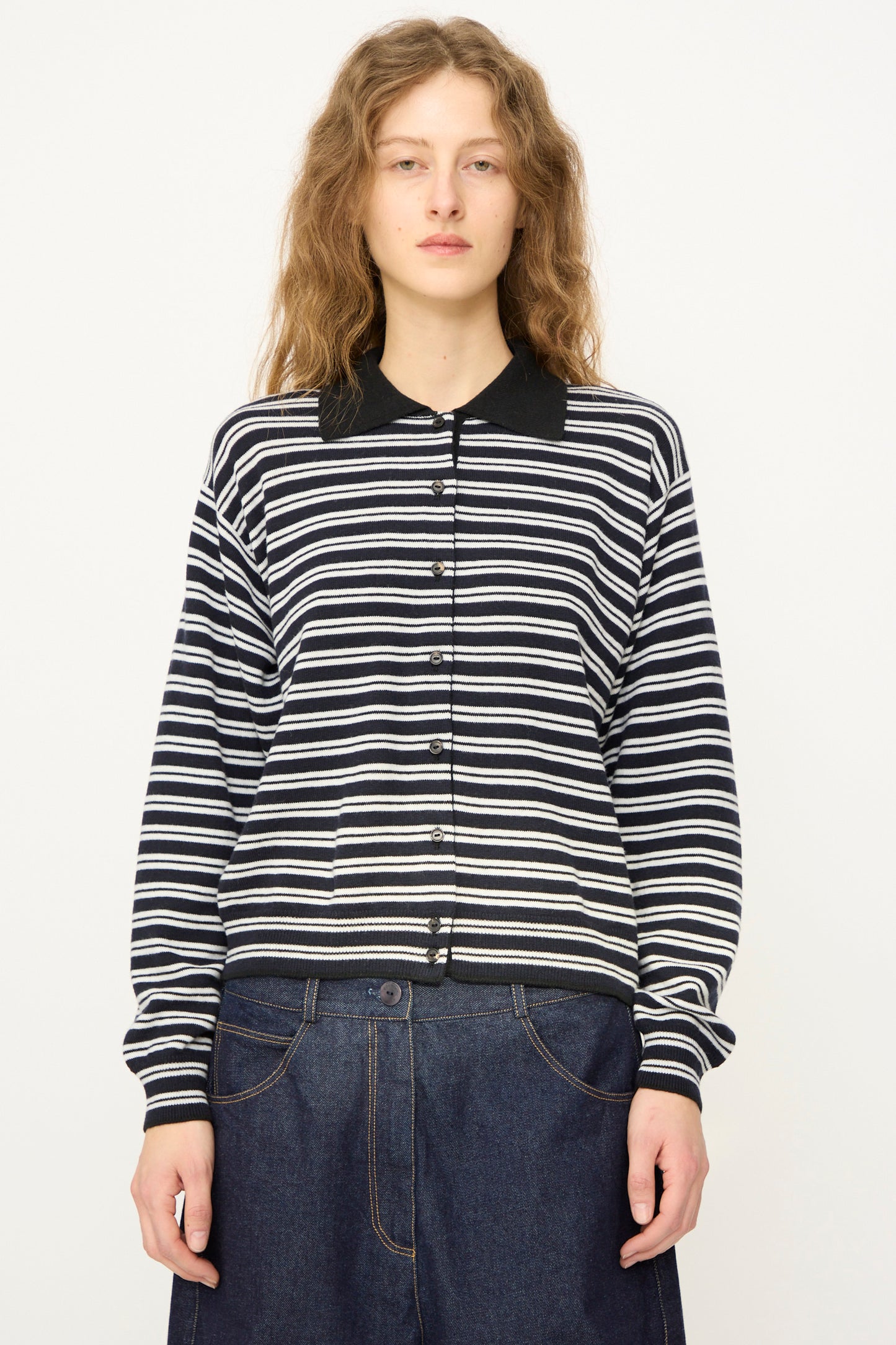 A person with wavy hair wears the Cordera Merino Wool Polo Cardigan in Striped, featuring a black collar, paired with wide-leg denim jeans, standing against a plain white background.