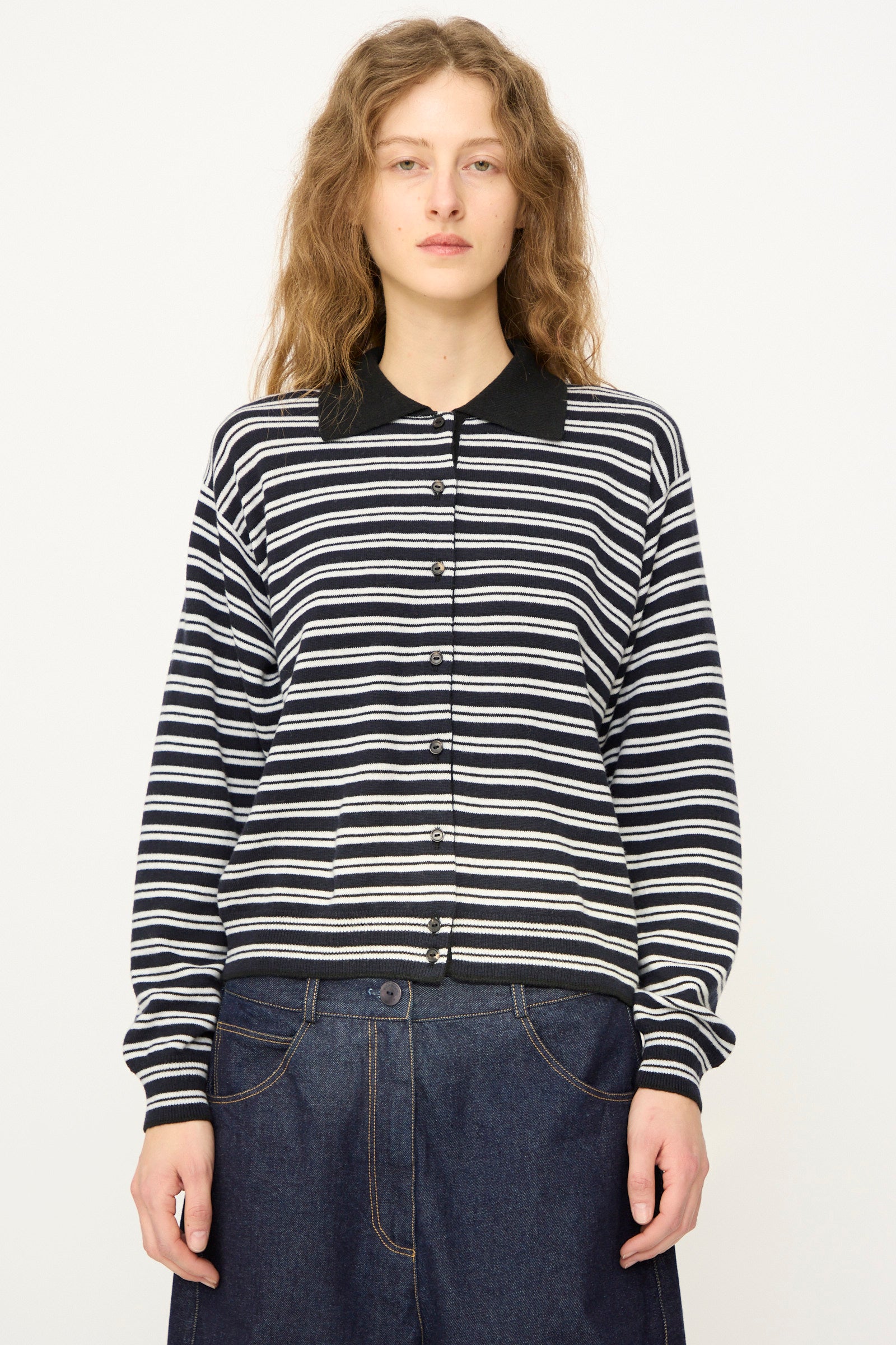 A person with wavy hair wears the Cordera Merino Wool Polo Cardigan in Striped, featuring a black collar, paired with wide-leg denim jeans, standing against a plain white background.