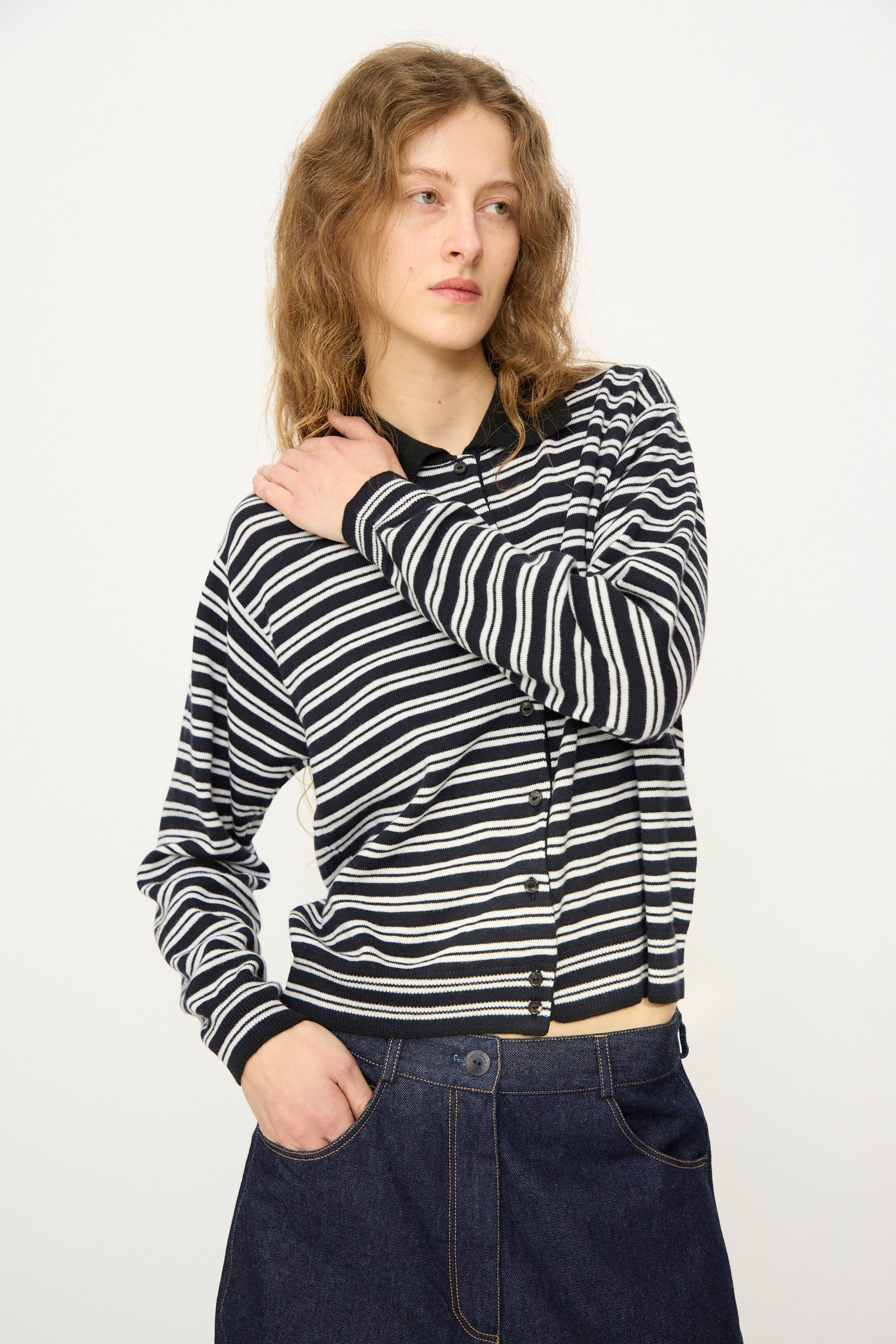 A person with wavy hair wears the Cordera Merino Wool Polo Cardigan in Striped with a dark denim skirt, standing against a plain light background and looking to the side.