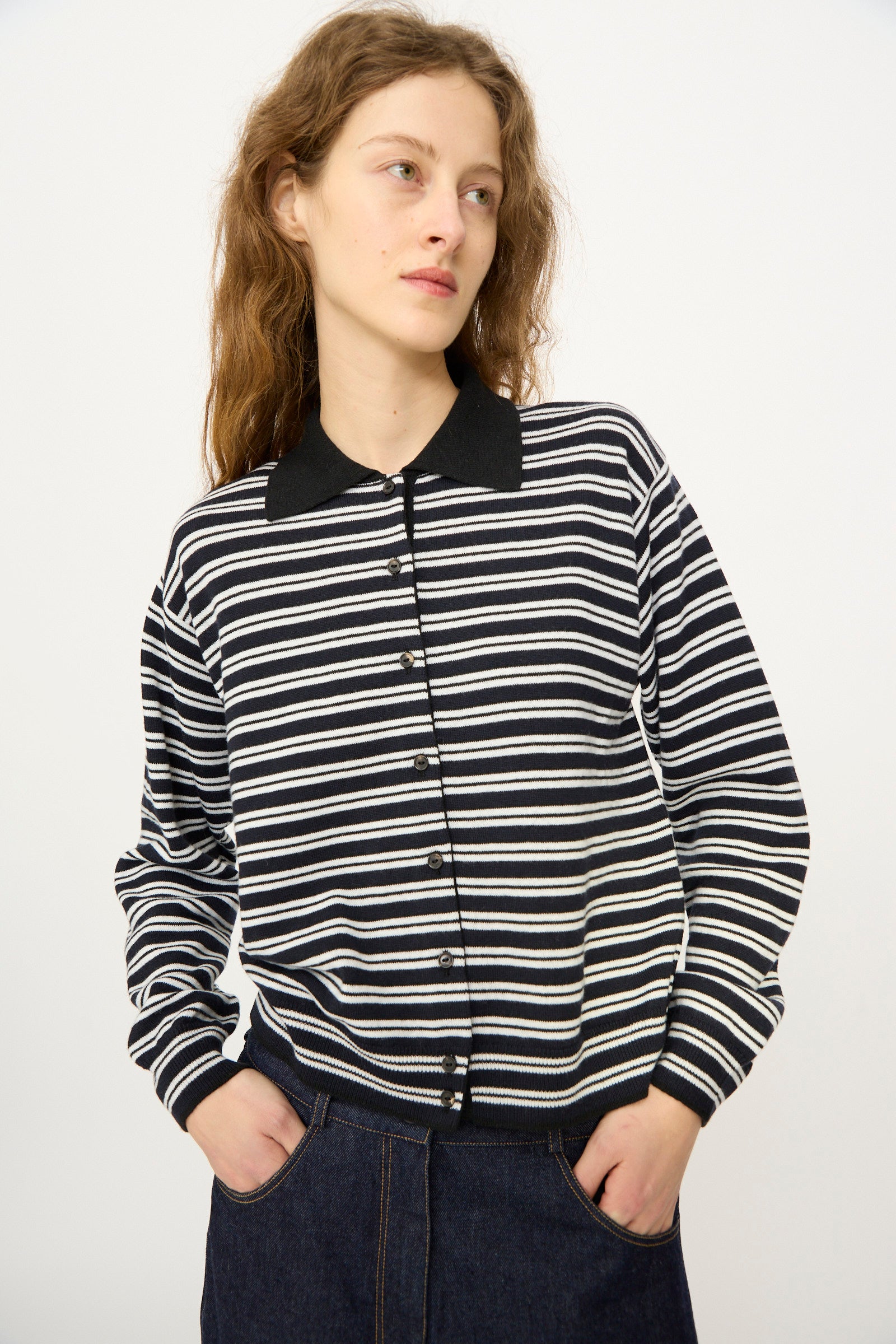 A person with long, wavy hair wears the Cordera Merino Wool Polo Cardigan in Striped with a black collar and dark jeans, standing against a plain background.