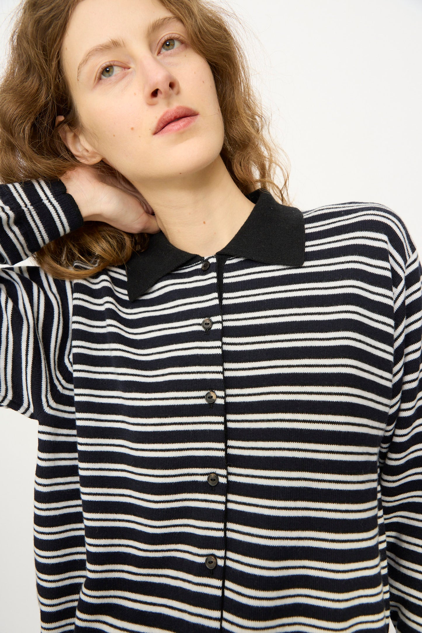 A person with wavy hair wears the Cordera Merino Wool Polo Cardigan in Striped, featuring black and white stripes and a black collar, while looking slightly upward with one hand behind their neck.