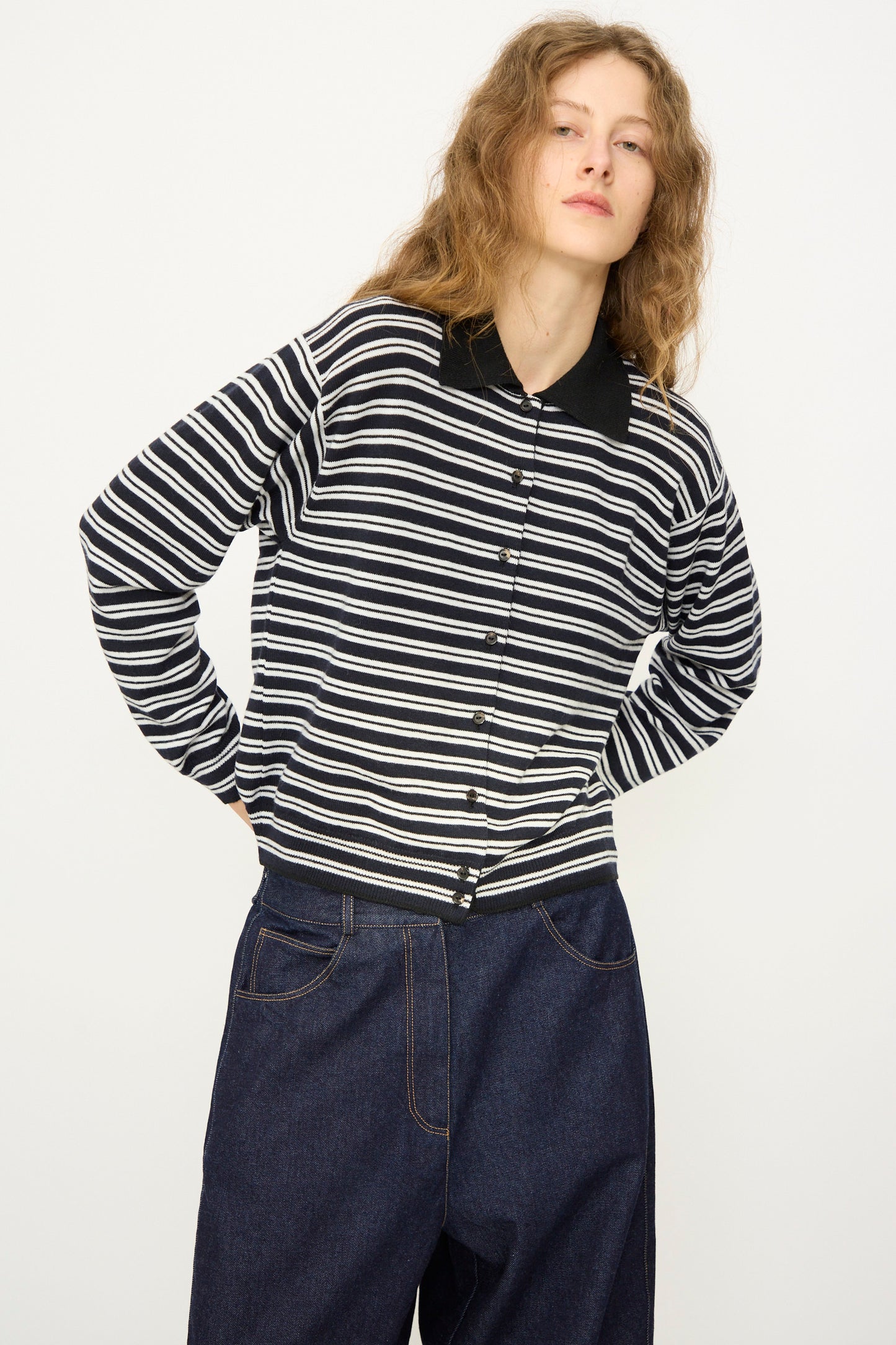 A person with long, wavy hair stands against a plain light background wearing Cordera's Merino Wool Polo Cardigan in Striped with a black collar and dark blue jeans.