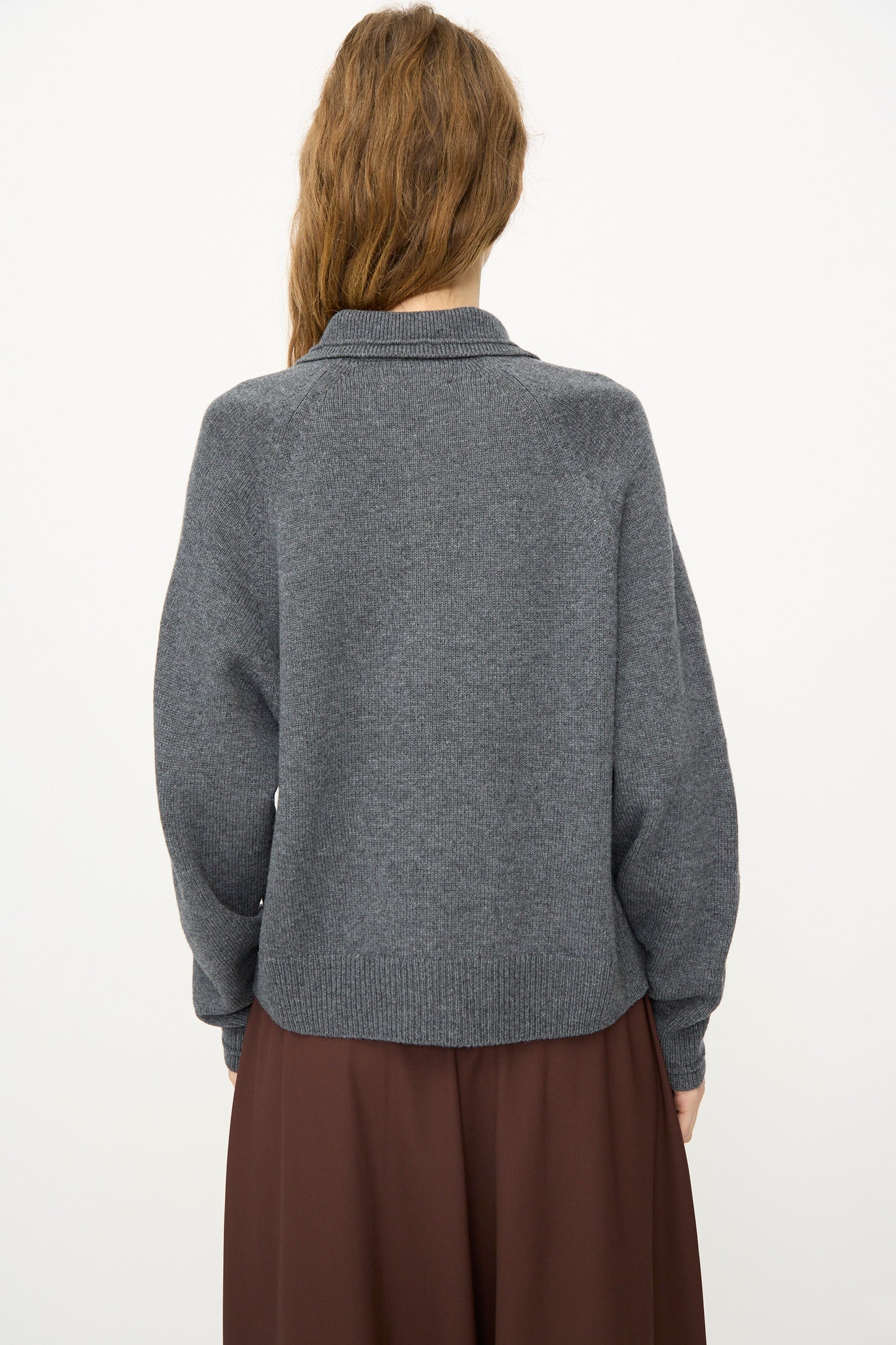 A person with long brown hair wears the Cordera Merino Wool Polo Sweater in Grey, paired with a brown skirt and photographed from the back against a plain white background.