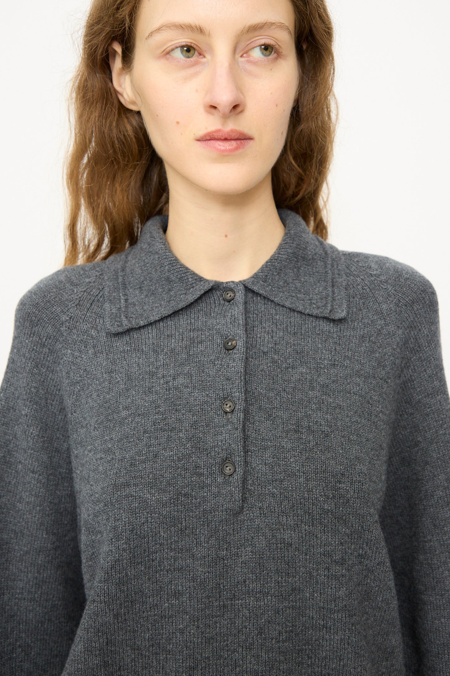 A person with long brown hair wears the Cordera Merino Wool Polo Sweater in Grey, featuring a relaxed fit and four buttons, facing slightly left against a plain background.