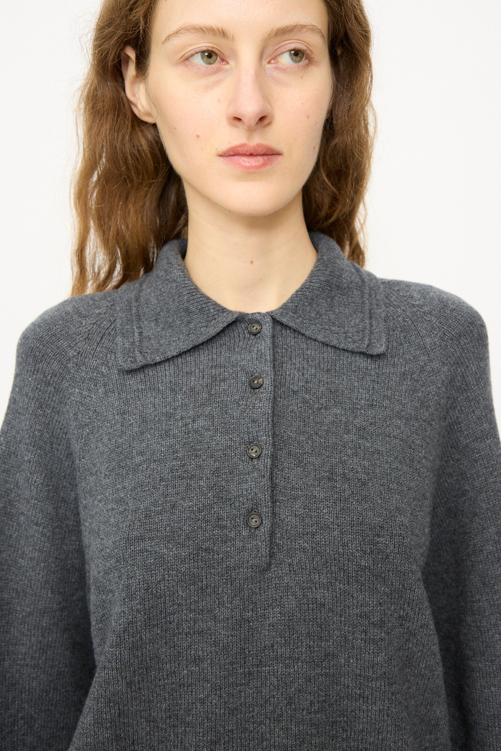 A person with long brown hair wears the Cordera Merino Wool Polo Sweater in Grey, featuring a relaxed fit and four buttons, facing slightly left against a plain background.