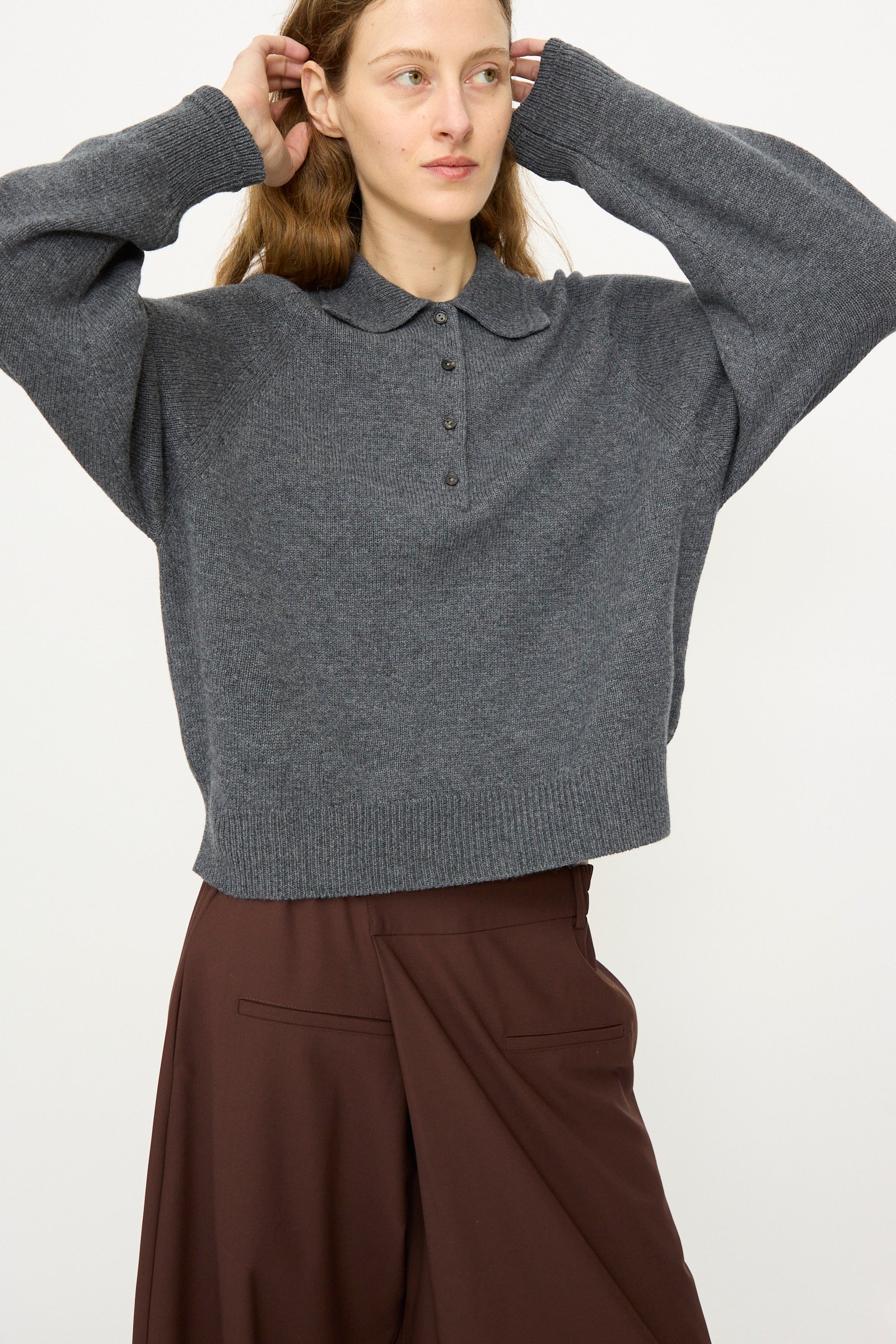 A person wears the Cordera Merino Wool Polo Sweater in Grey, a relaxed fit knit with a collar, paired with brown wide-leg pants, standing against a plain background with hands touching their head.
