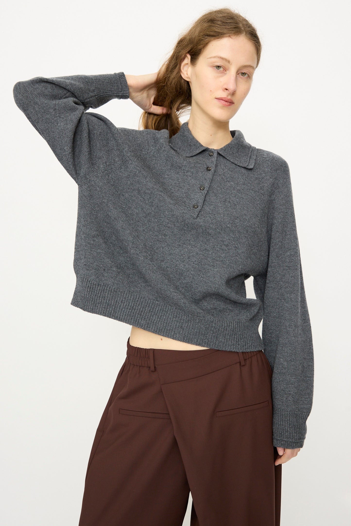 A person with long hair holds the back of their head, wearing Cordera’s Merino Wool Polo Sweater in Grey and brown pants, standing against a plain light background.