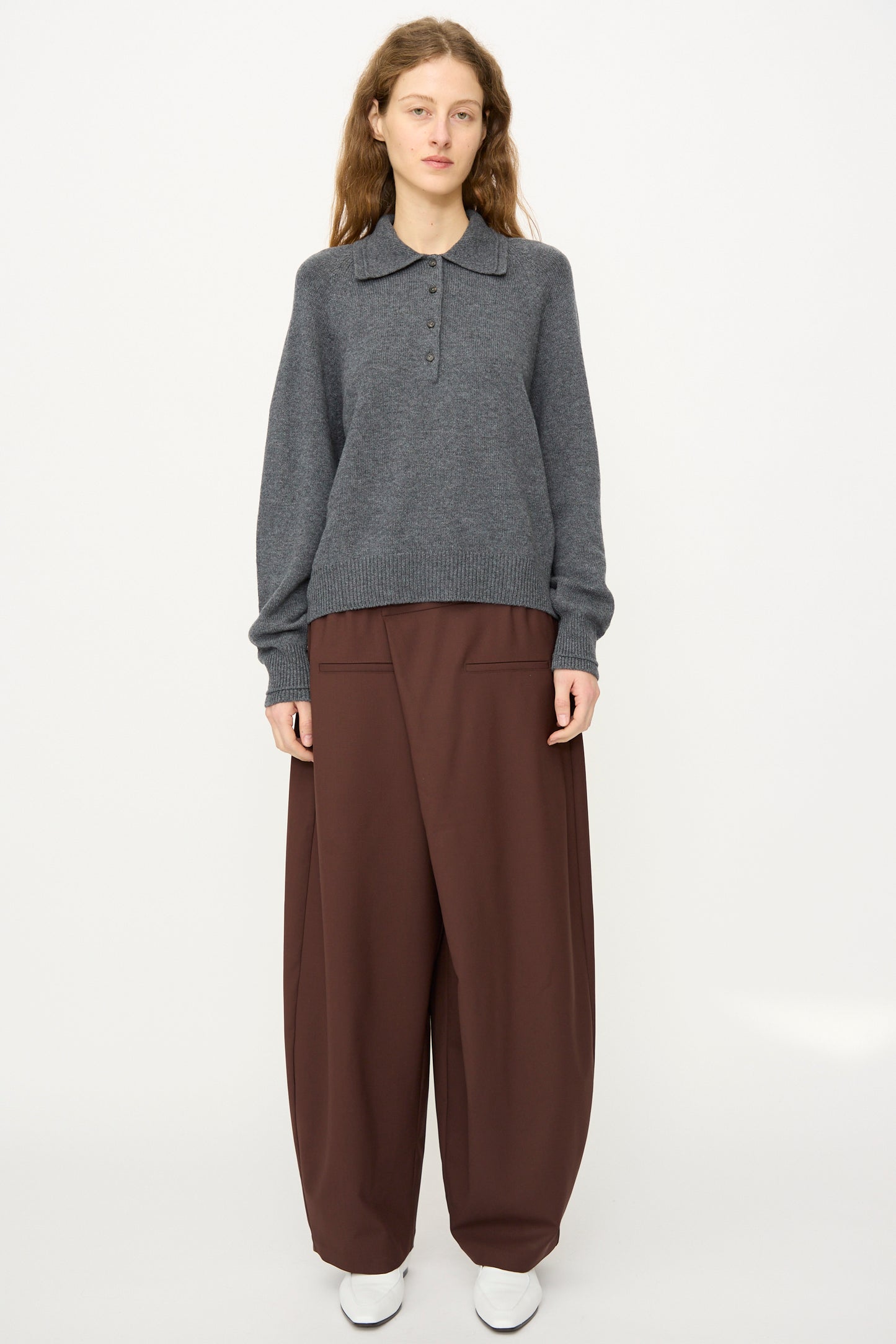 A person faces forward against a plain white background, wearing Cordera's Merino Wool Polo Sweater in Grey, styled with wide brown trousers and white shoes.