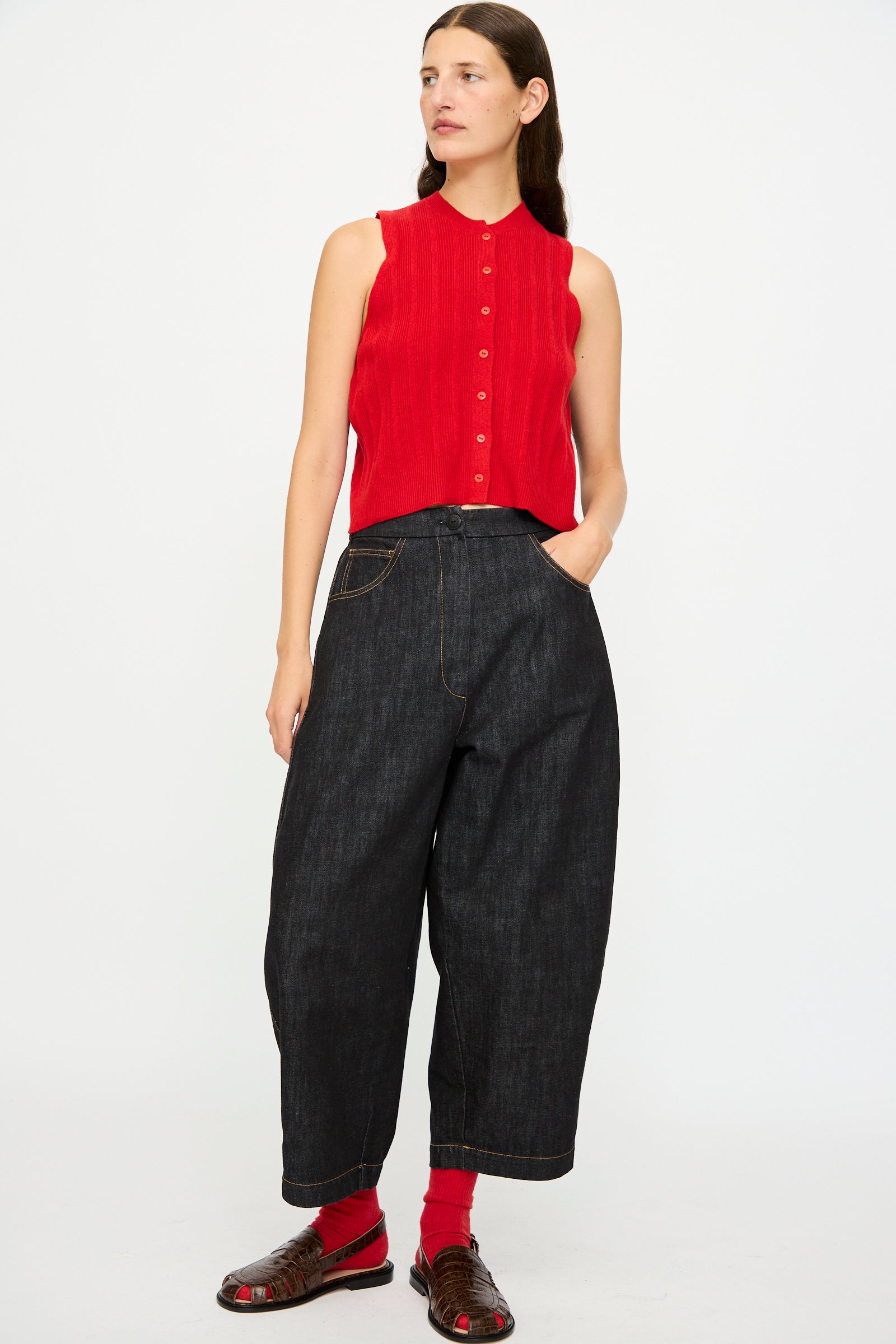A woman poses against a plain background wearing the Cordera Merino Wool Ribbed Waistcoat in Red, loose dark denim pants, red socks, and brown sandals with one hand in her pocket.