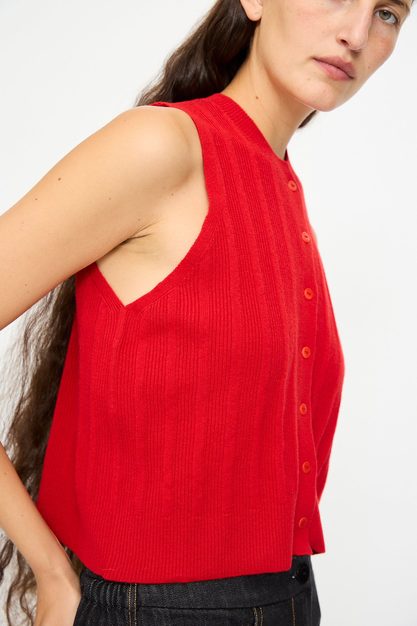 A woman, shown from the side against a plain background, wears Cordera’s Merino Wool Ribbed Waistcoat in Red styled as a cropped sleeveless top with black pants.