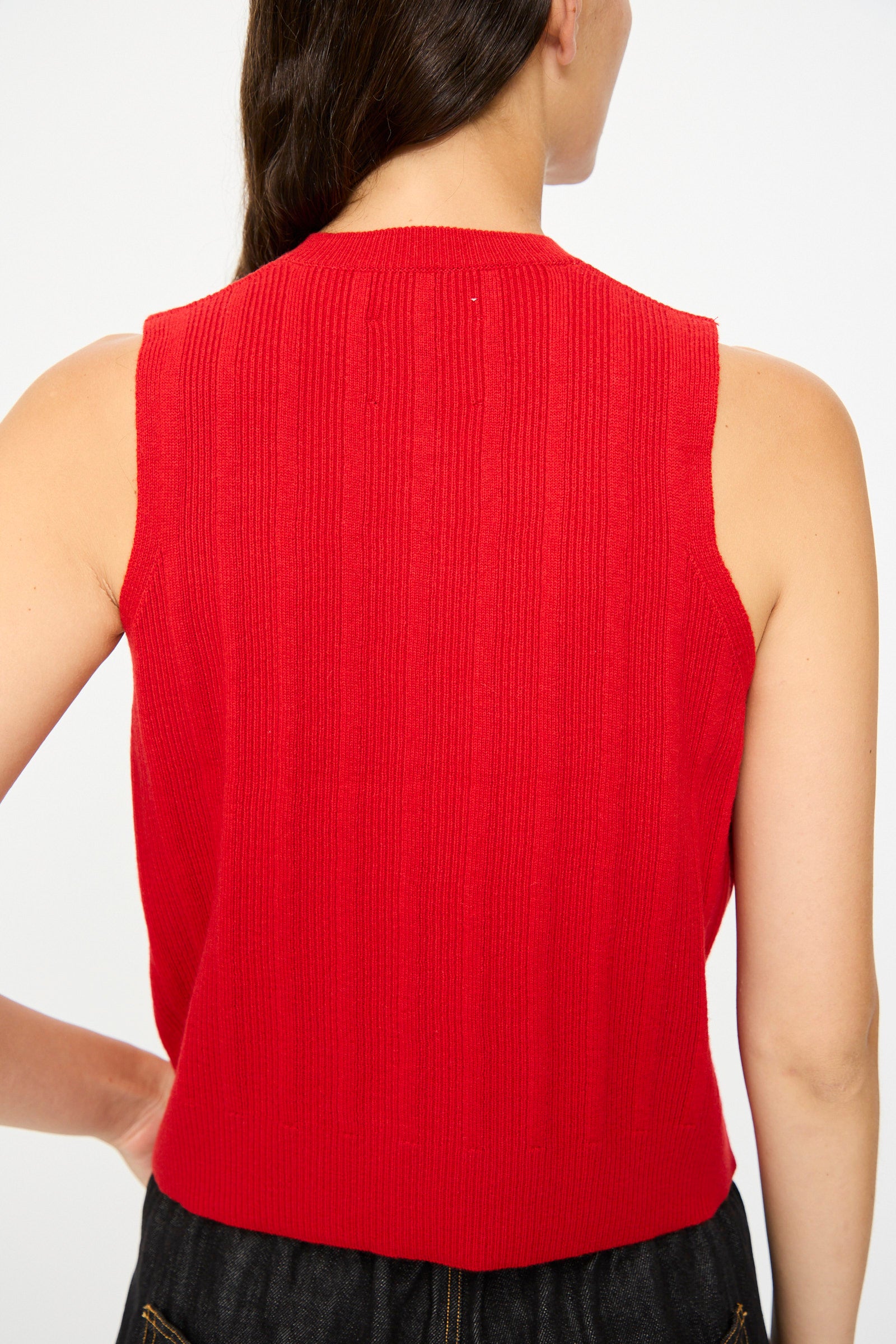 A woman with long dark hair is seen from behind wearing Cordera's Merino Wool Ribbed Waistcoat in Red, a sleeveless piece made in Spain, paired with dark pants.