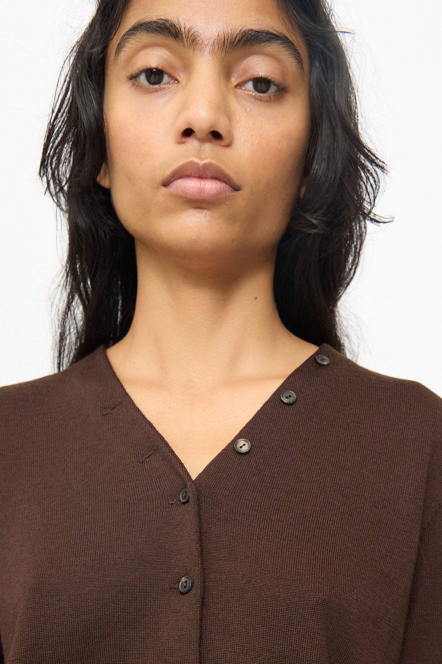 A person with long dark hair wears the Cordera Merino Wool Button Cardigan in Brown, featuring an asymmetrical button detail and photographed from the chest up against a plain background.