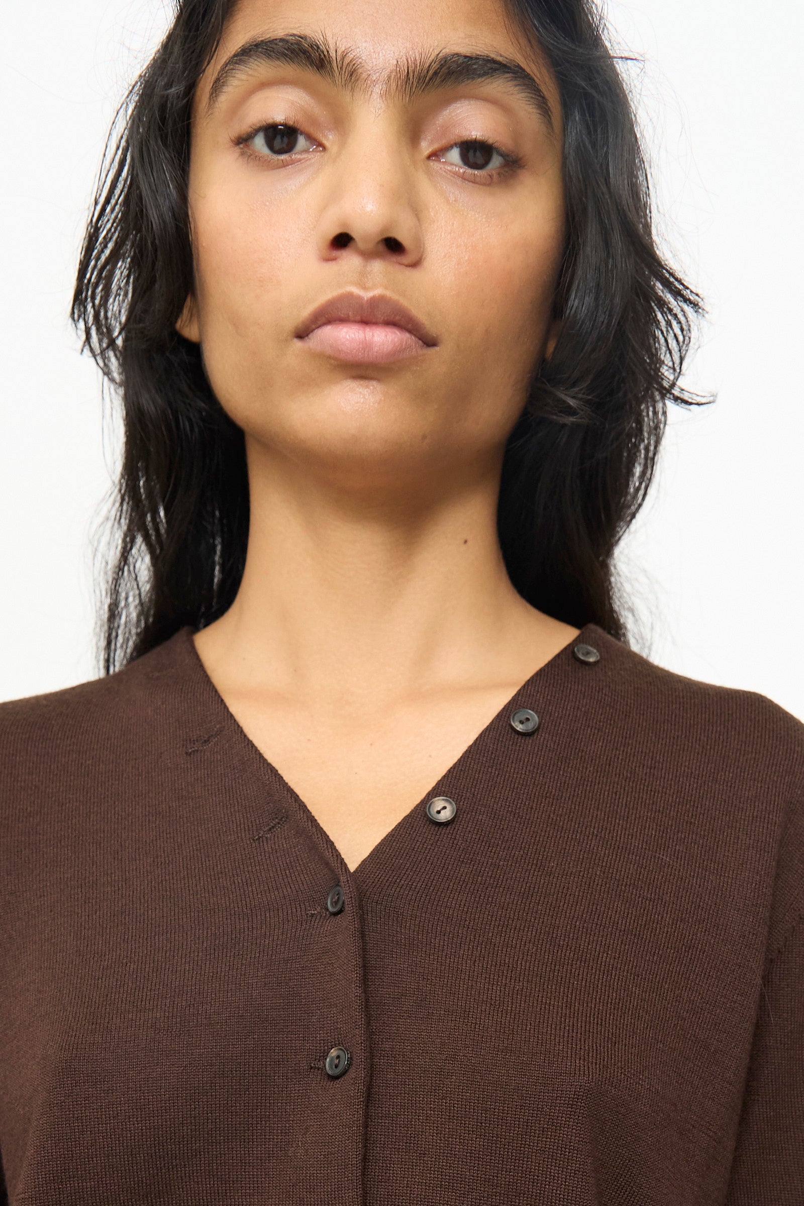 A person with long dark hair wears the Cordera Merino Wool Button Cardigan in Brown, featuring an asymmetrical button detail and photographed from the chest up against a plain background.