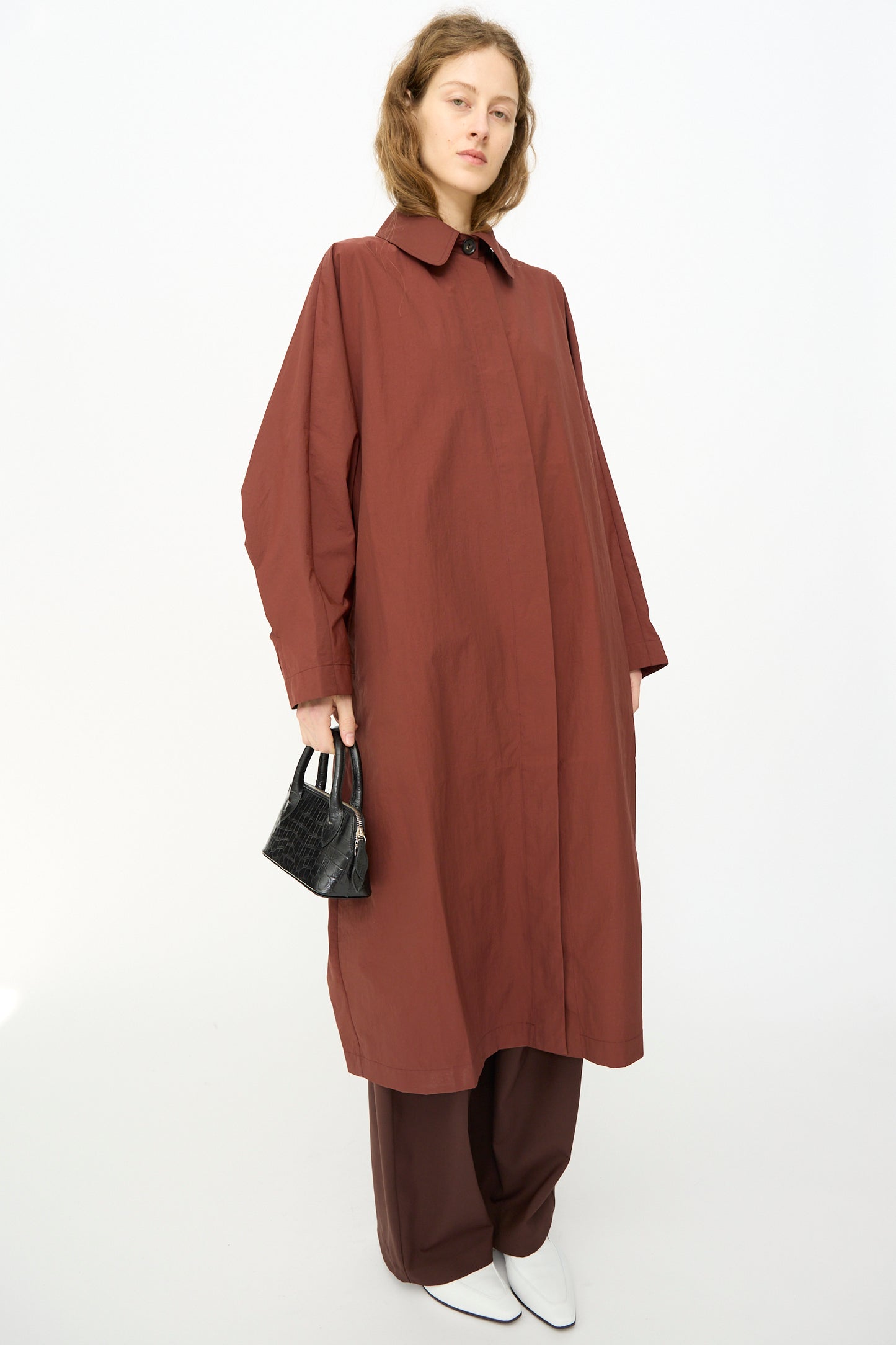 A person wearing a loose-fitting, long maroon coat over brown pants holds the Cordera Mini Bag Crocodile in Black, standing against a plain white background.