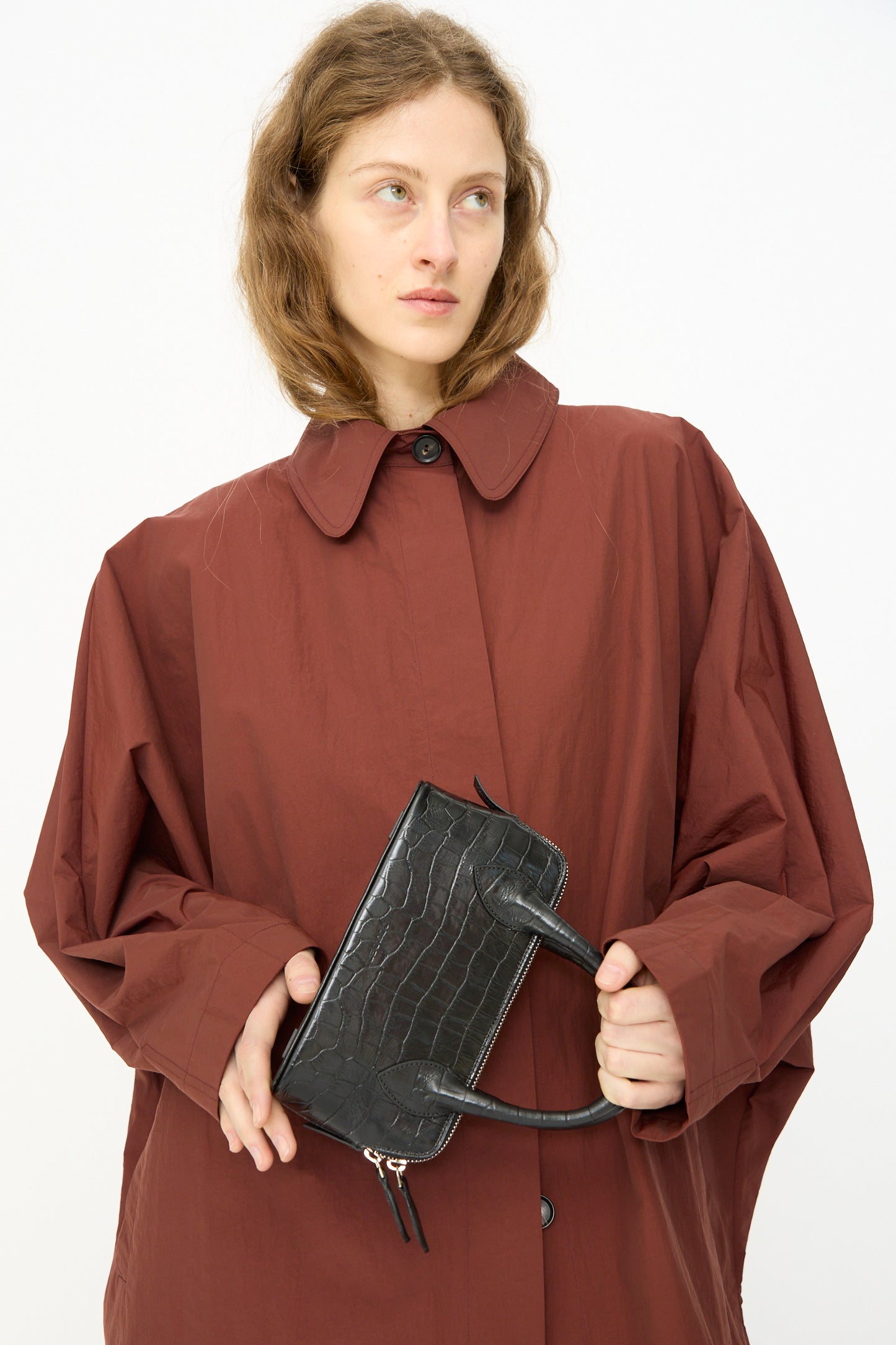 A person in an oversized brown coat holds the Cordera Mini Bag Crocodile in Black, a small rectangular calfskin leather handbag, with both hands against a plain white background.