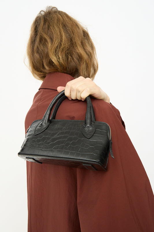 A person with wavy brown hair in a brown coat holds the Cordera Mini Bag Crocodile in Black over their shoulder, facing away from the camera.