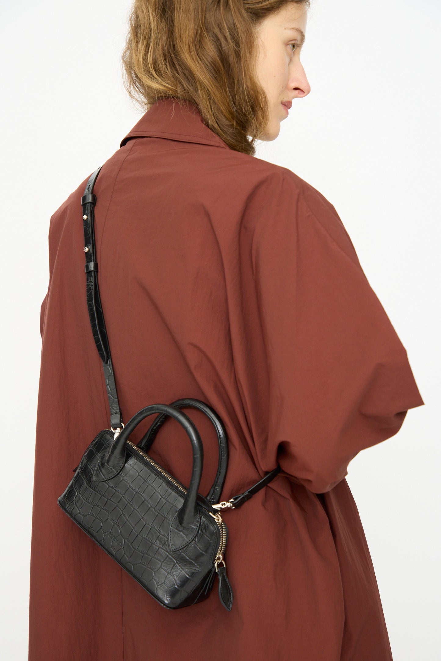 A person wears a loose brown coat with the Cordera Mini Bag Crocodile in Black, a mini rectangular crossbody crafted from black crocodile-texture leather, slung diagonally across their back.