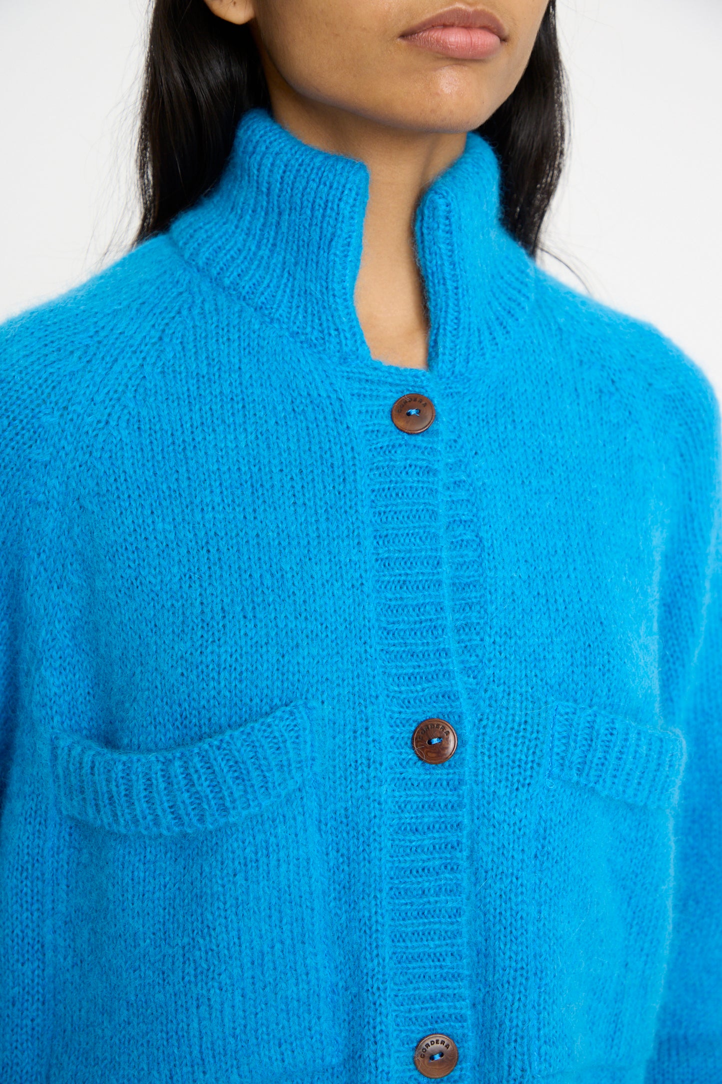 A close-up of a person wearing the Cordera Mohair and Baby Alpaca Polo Jacket in Cerúleo, a bright blue high-collared knit featuring wooden buttons and a cozy mohair blend.