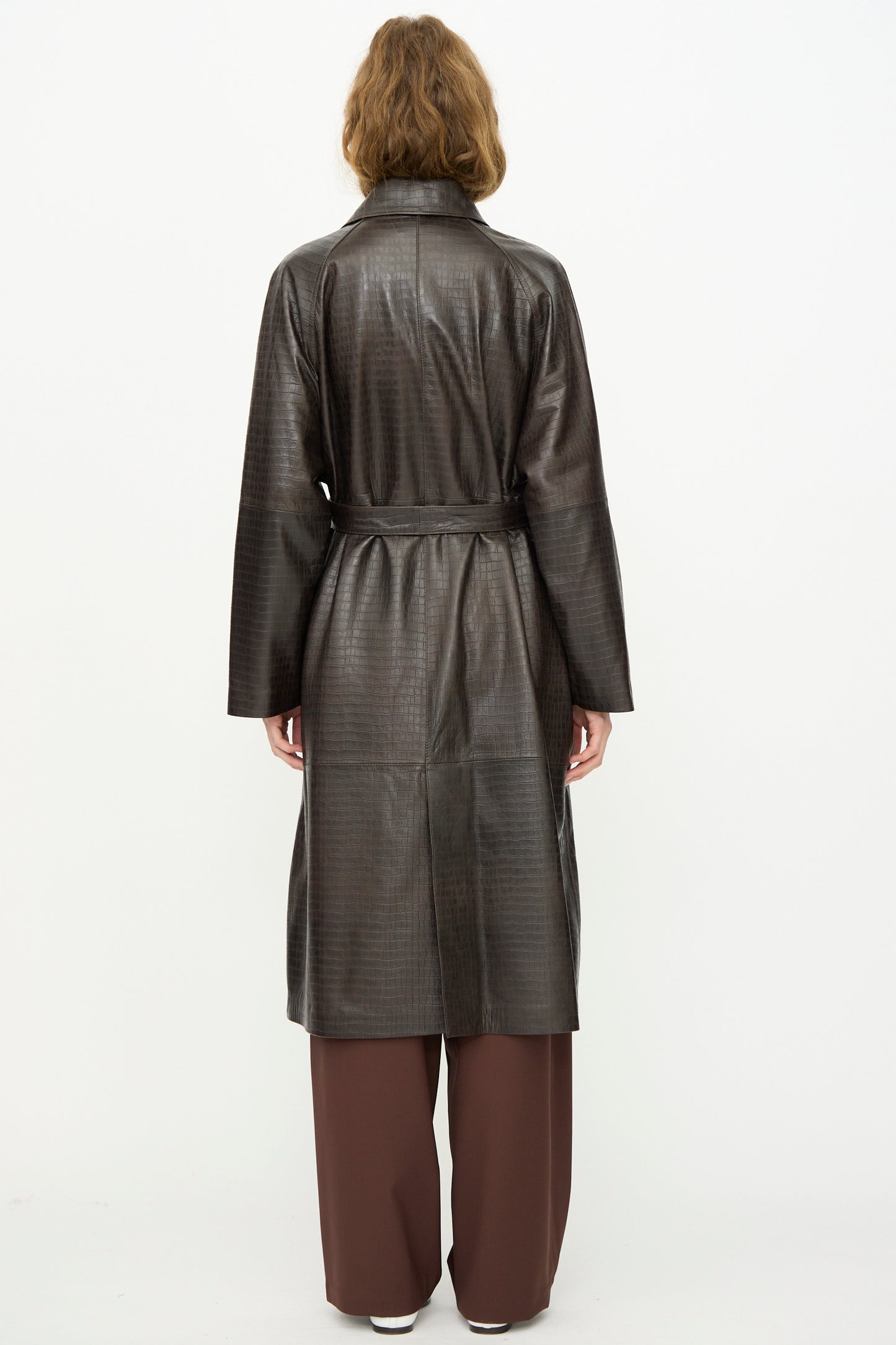 A person stands facing away in the Cordera Nappa Long Coat in Crocodile, a long dark brown embossed coat with a belt, styled over brown pants and white shoes against a plain white background.