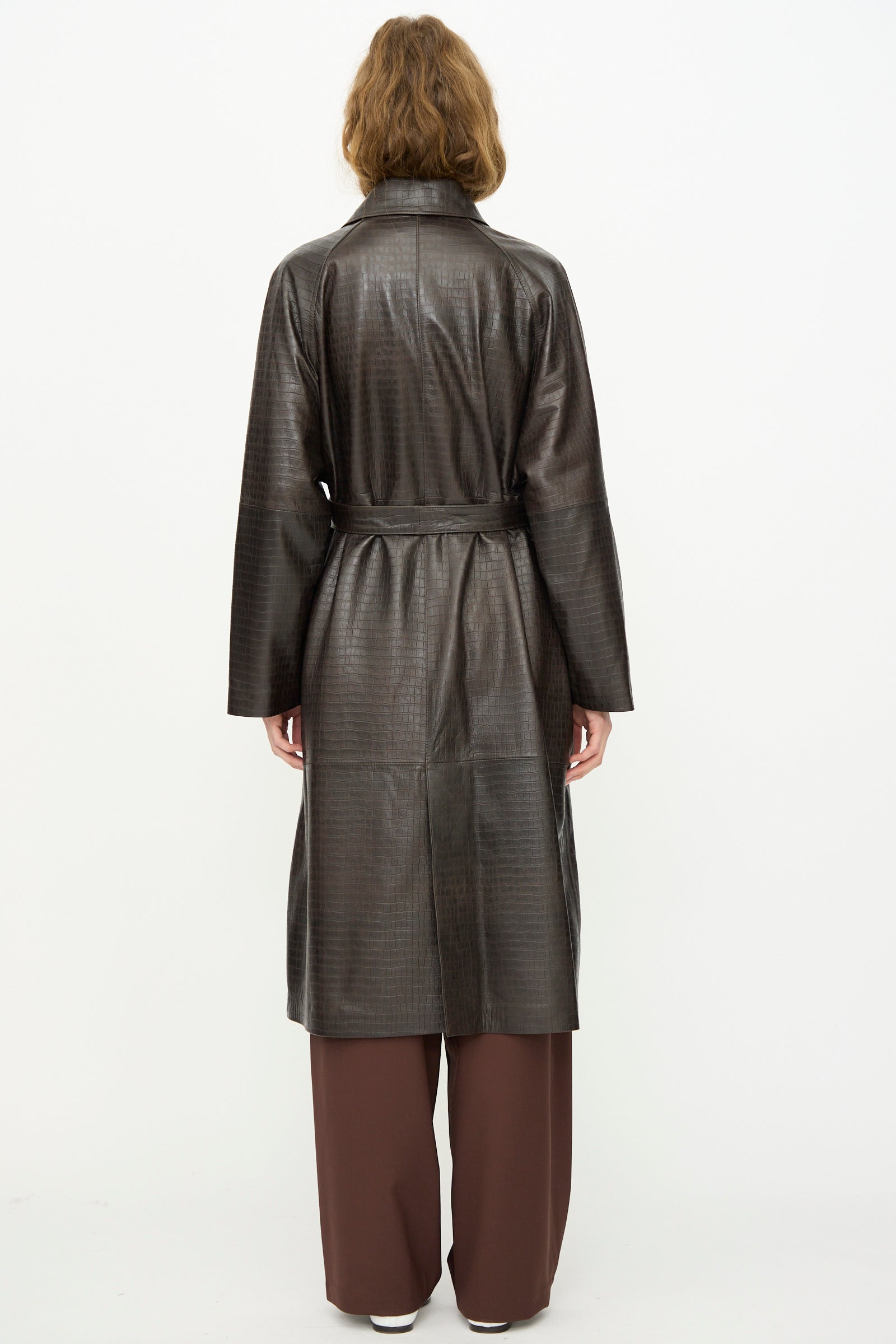 A person stands facing away in the Cordera Nappa Long Coat in Crocodile, a long dark brown embossed coat with a belt, styled over brown pants and white shoes against a plain white background.