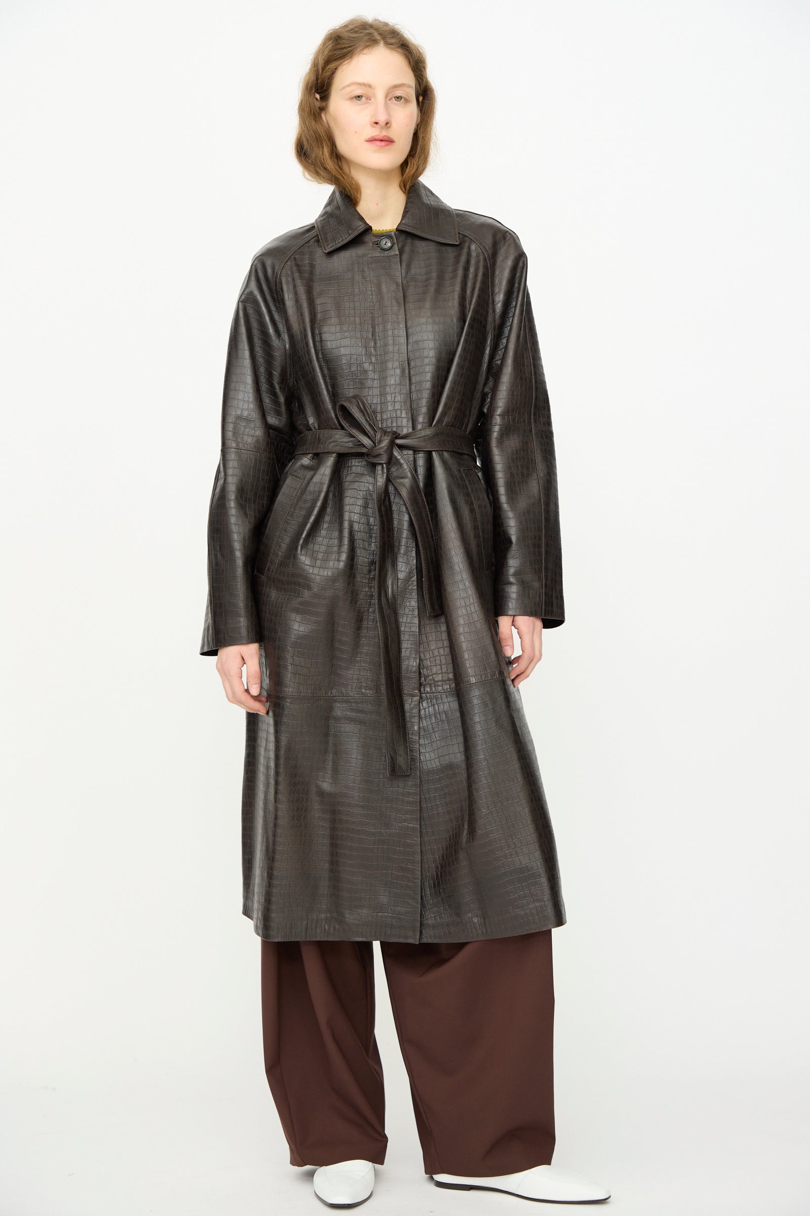 A person wears the Cordera Nappa Long Coat in Crocodile over wide-legged brown pants and white shoes, standing against a plain white background.