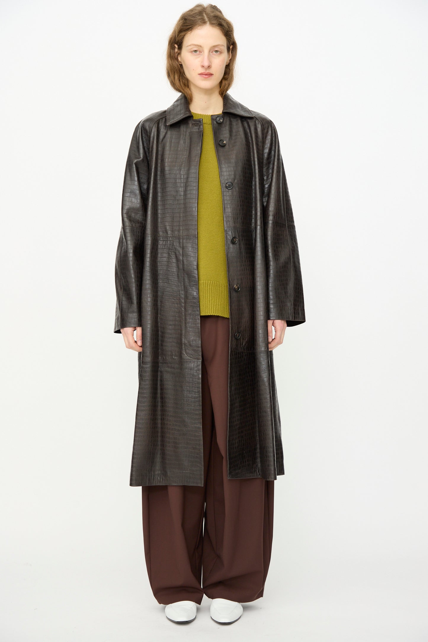 A person faces forward against a white background, wearing Cordera's Nappa Long Coat in Crocodile over an olive green sweater, wide brown pants, and white shoes.