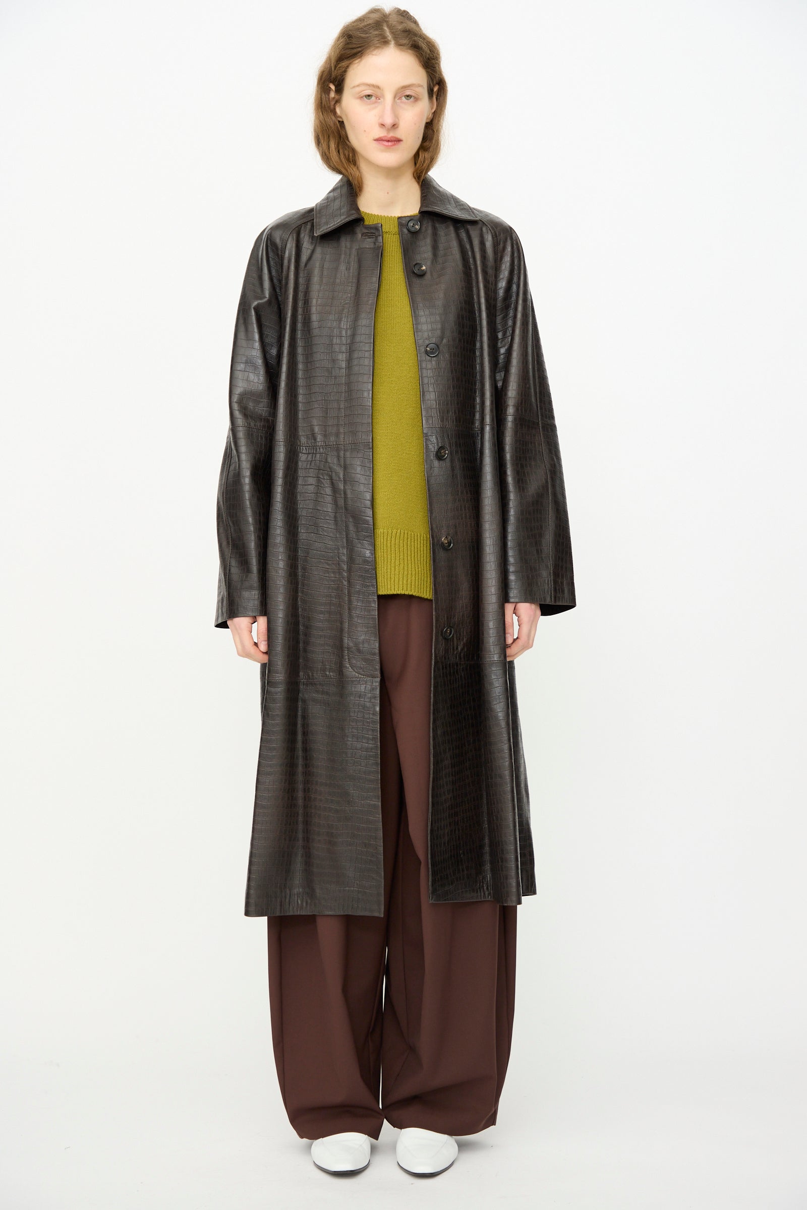 A person faces forward against a white background, wearing Cordera's Nappa Long Coat in Crocodile over an olive green sweater, wide brown pants, and white shoes.