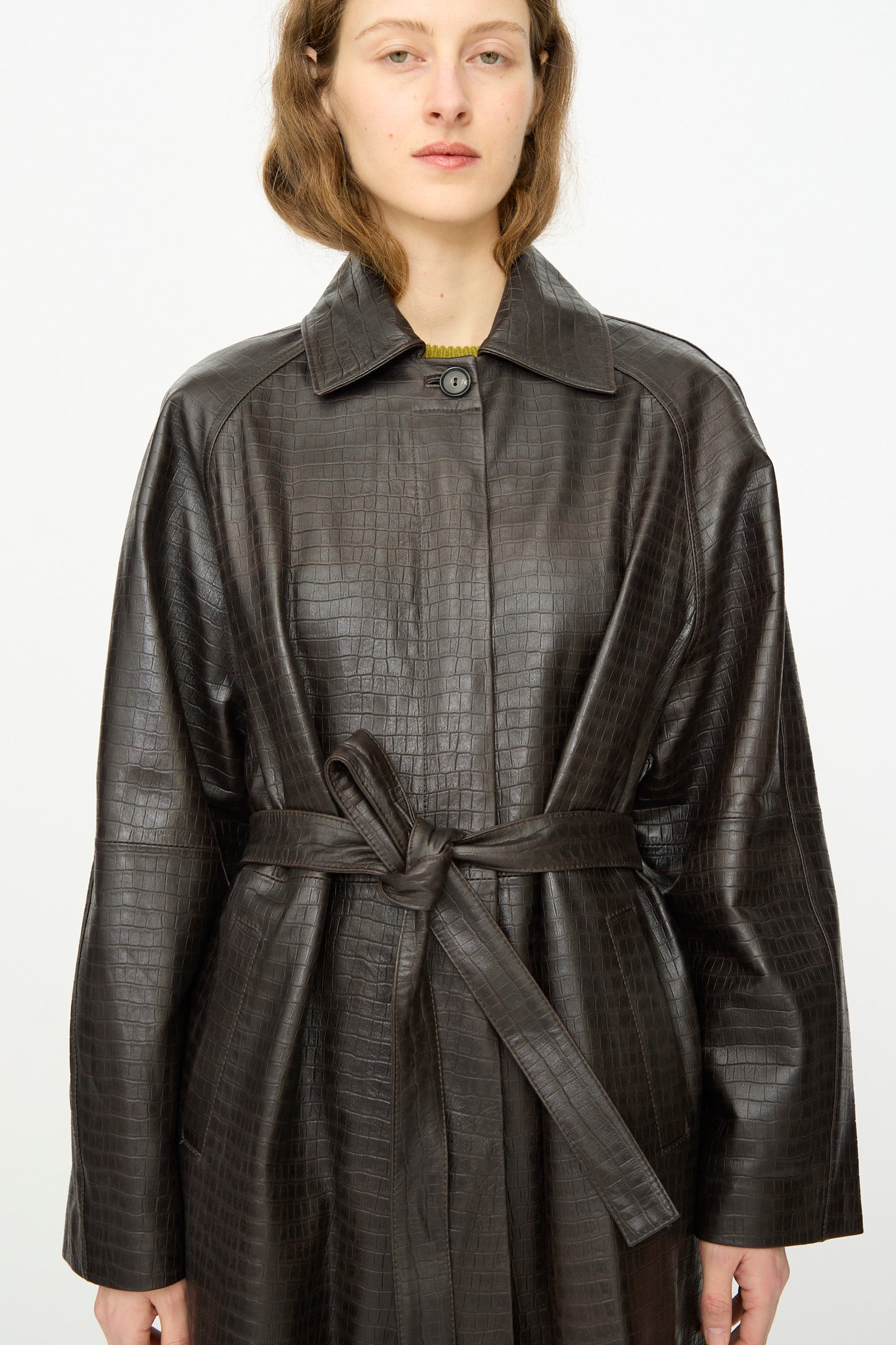 Person wearing the Cordera Nappa Long Coat in Crocodile, a dark brown, crocodile-embossed trench coat with a belt, standing against a plain white background.