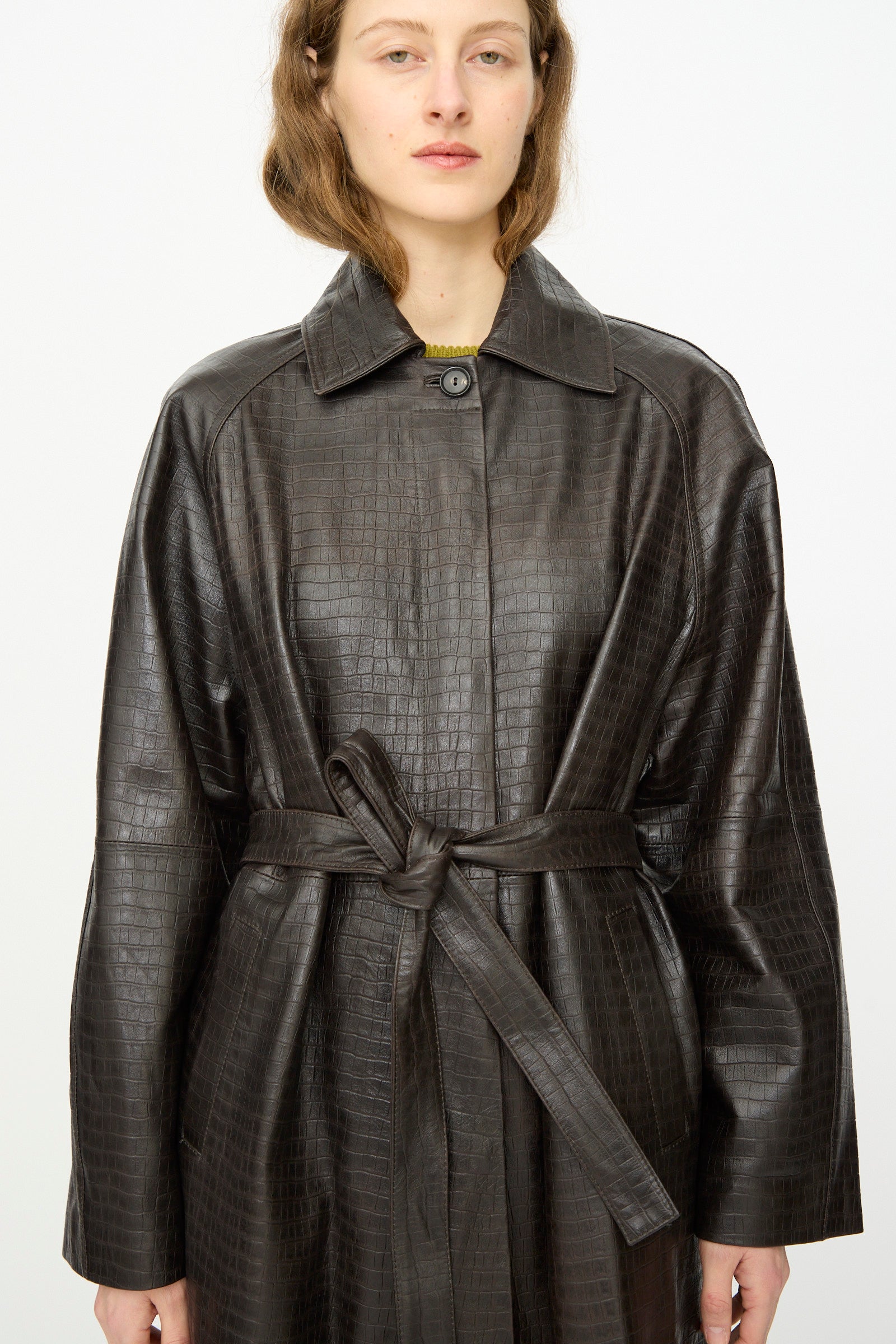 Person wearing the Cordera Nappa Long Coat in Crocodile, a dark brown, crocodile-embossed trench coat with a belt, standing against a plain white background.