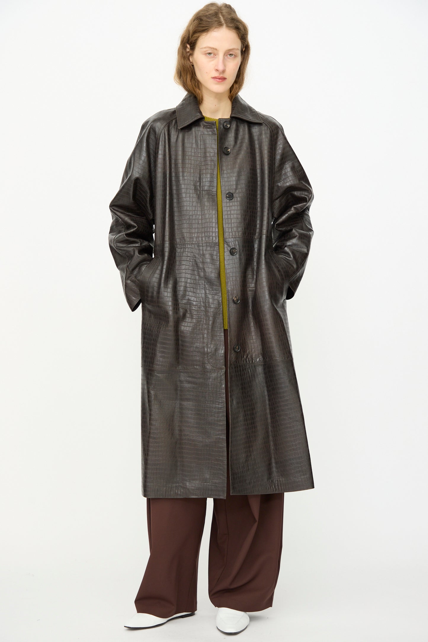 A person wears the Cordera Nappa Long Coat in Crocodile over a green top and brown wide-leg pants, posing against a plain white background.