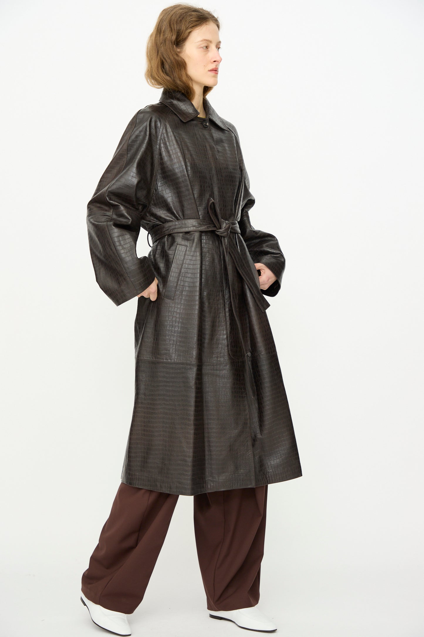 A person wears Cordera's Nappa Long Coat in Crocodile over wide-legged brown pants and white shoes, standing against a plain white background.