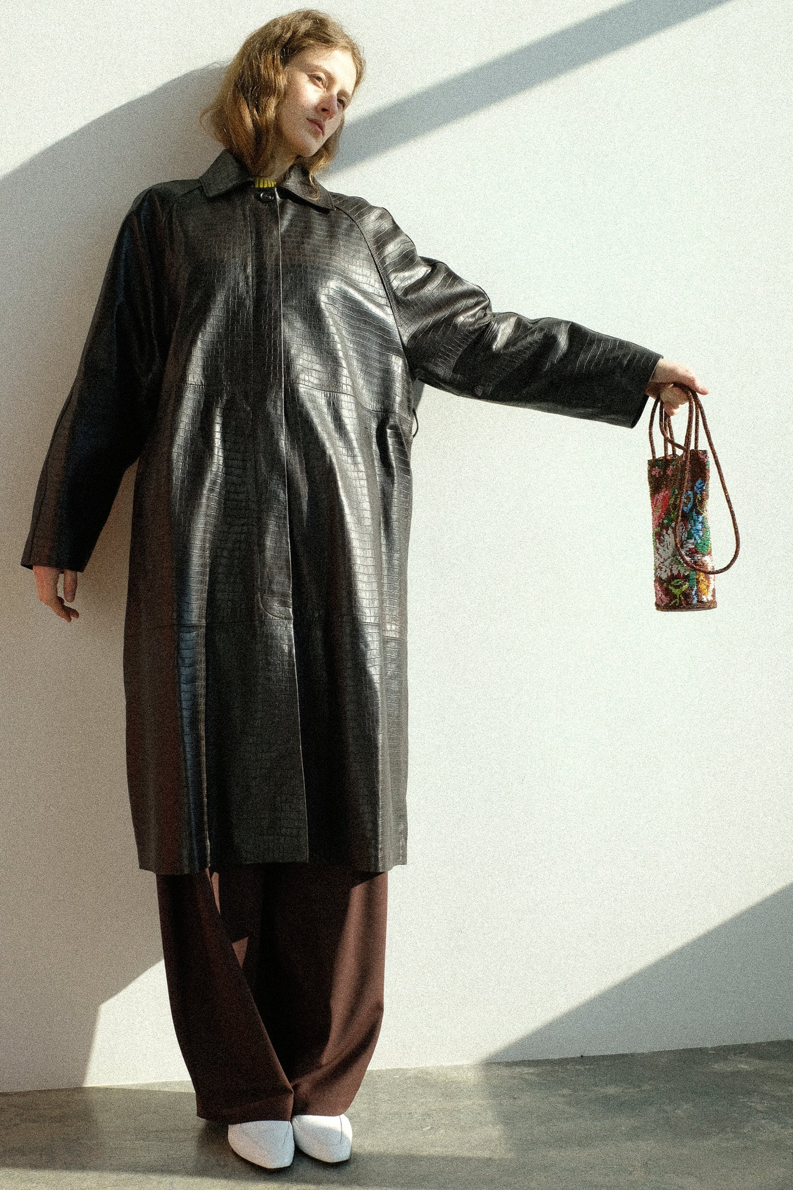A woman wearing Cordera's Nappa Long Coat in Crocodile, holding the Handmade Beaded Bag in Peonia against a white wall.