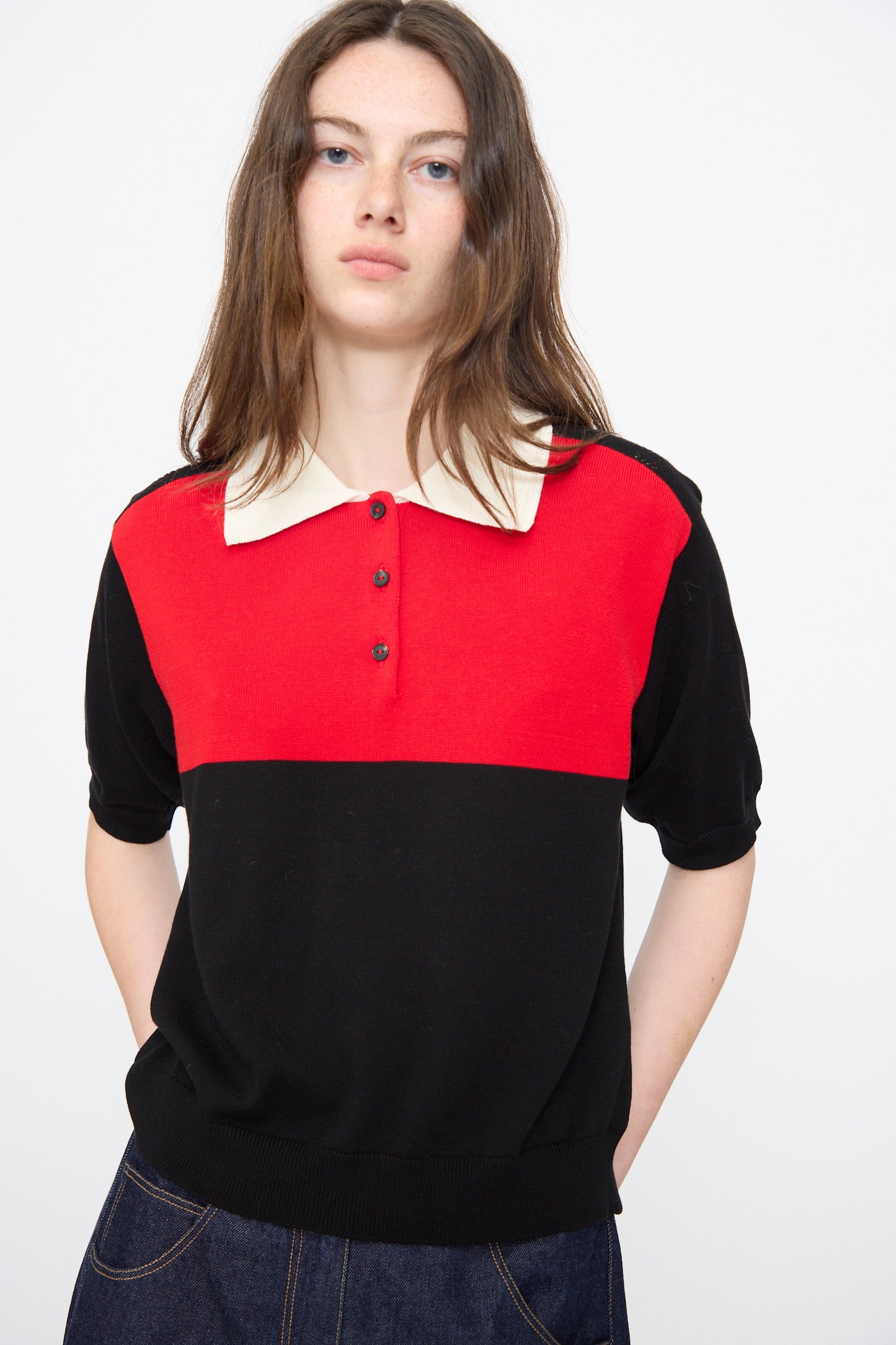 A woman with long brown hair wears the Cordera Cotton Polo in Colorblock, featuring a red upper section, black lower section, and cream collar, paired with dark denim jeans.