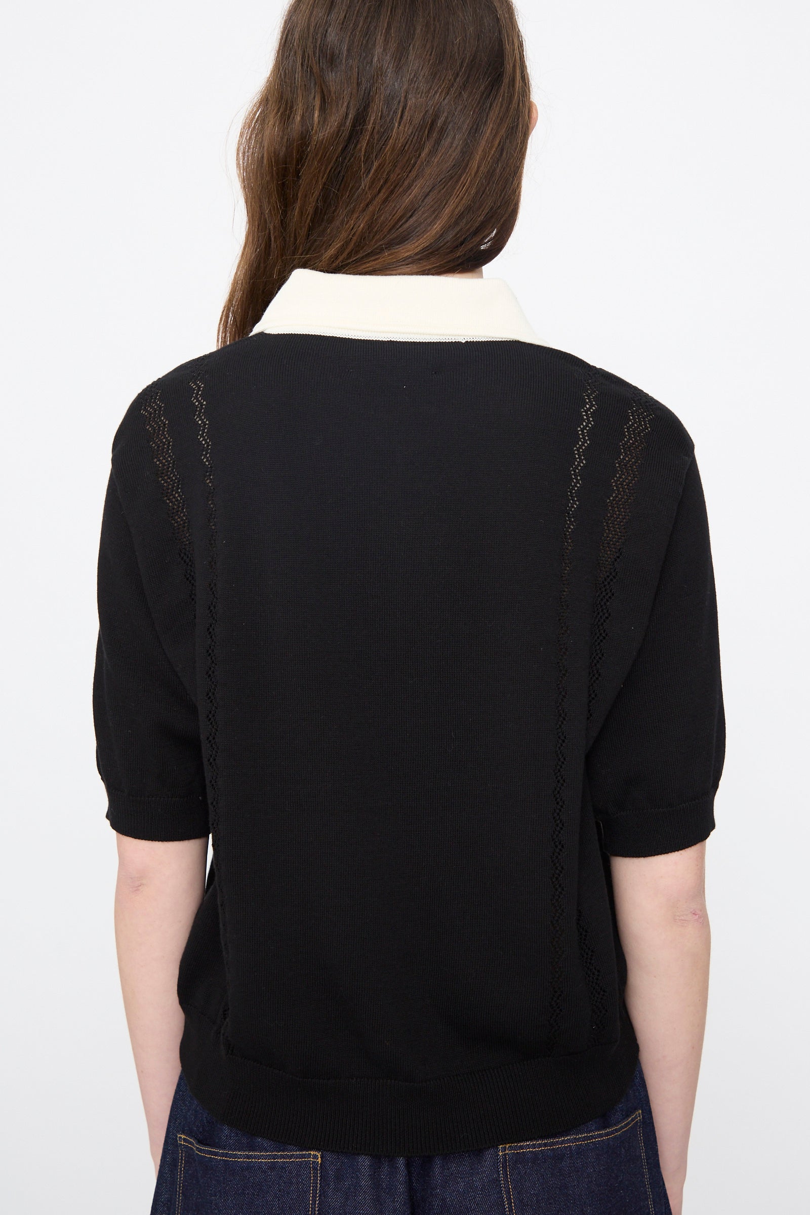Person with long brown hair faces away, wearing the Cordera Cotton Polo in Colorblock— a relaxed fit black knit cotton shirt with a white collar—paired with dark blue pants, set against a plain light background.
