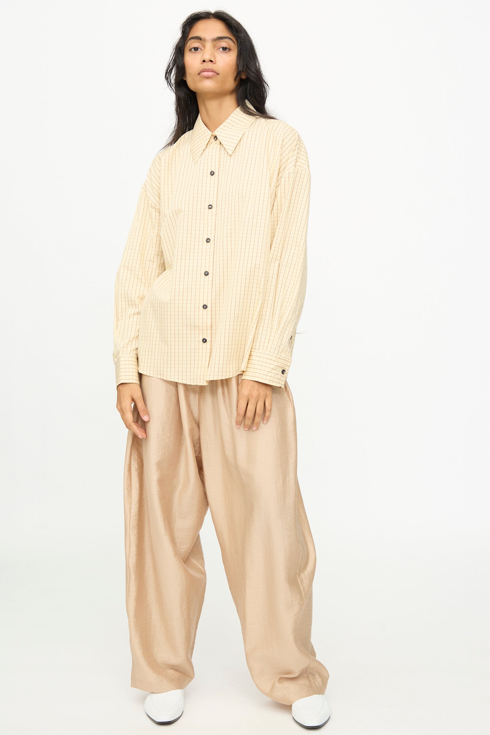 A person stands against a plain background wearing the Cordera Oversized Shirt in Checked, paired with wide light tan pants and white shoes, with a neutral facial expression.