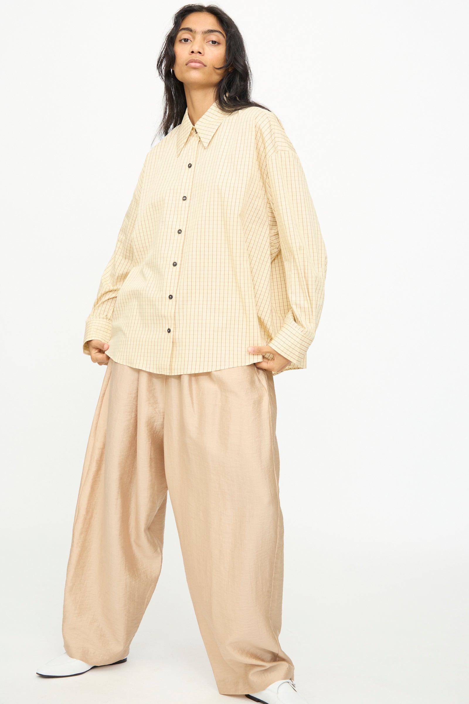 A woman wearing the Cordera Oversized Shirt in Checked stands against a plain white background, paired with wide-legged tan trousers, looking at the camera.