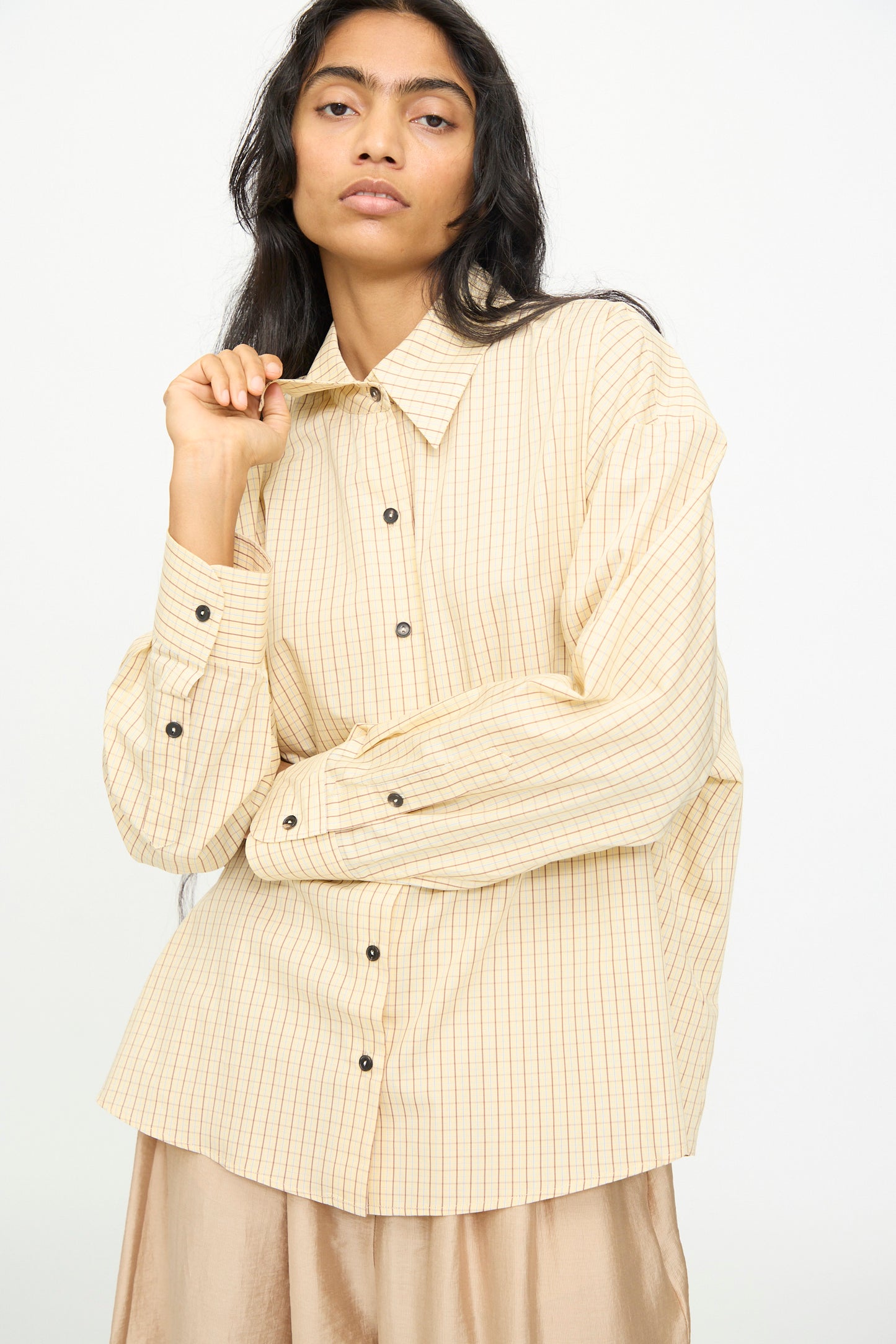 A woman poses against a plain background wearing the Cordera Oversized Shirt in Checked—an oversized, beige cotton button-up with black buttons and an Italian collar—paired with a light, shiny beige skirt.
