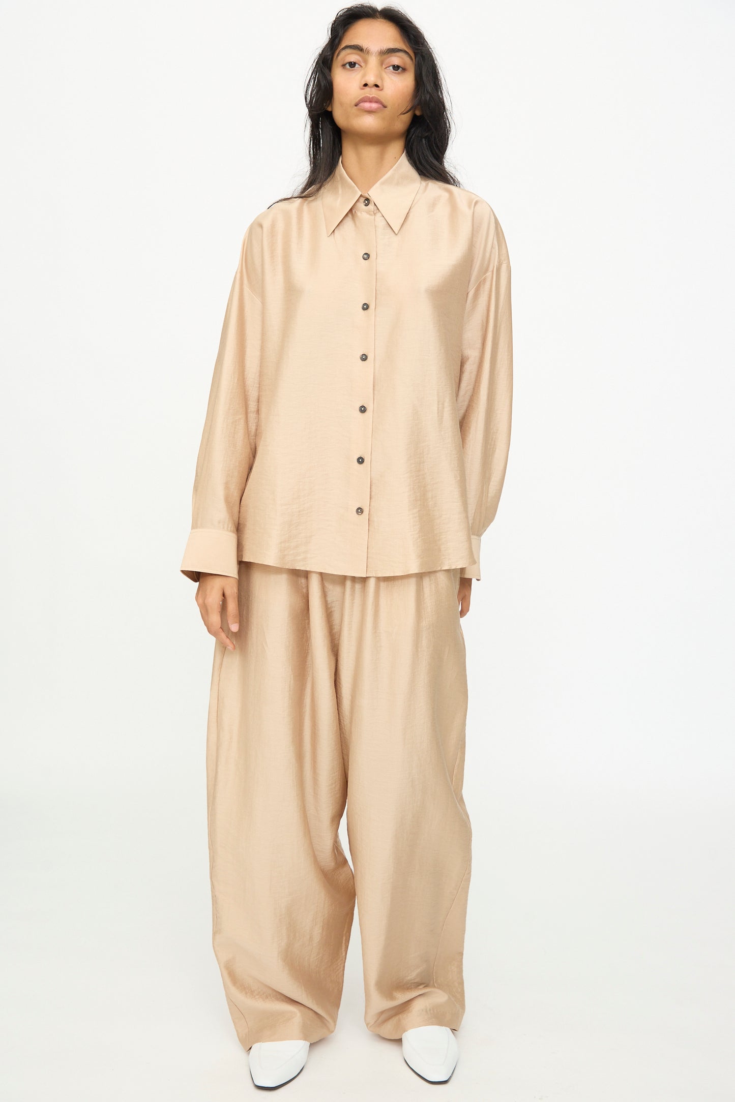 A person stands facing forward against a plain white background, wearing the Cordera Oversized Shirt in Beige, a loose-fitting lyocell blend with an Italian collar, paired with matching wide-leg pants and white shoes.