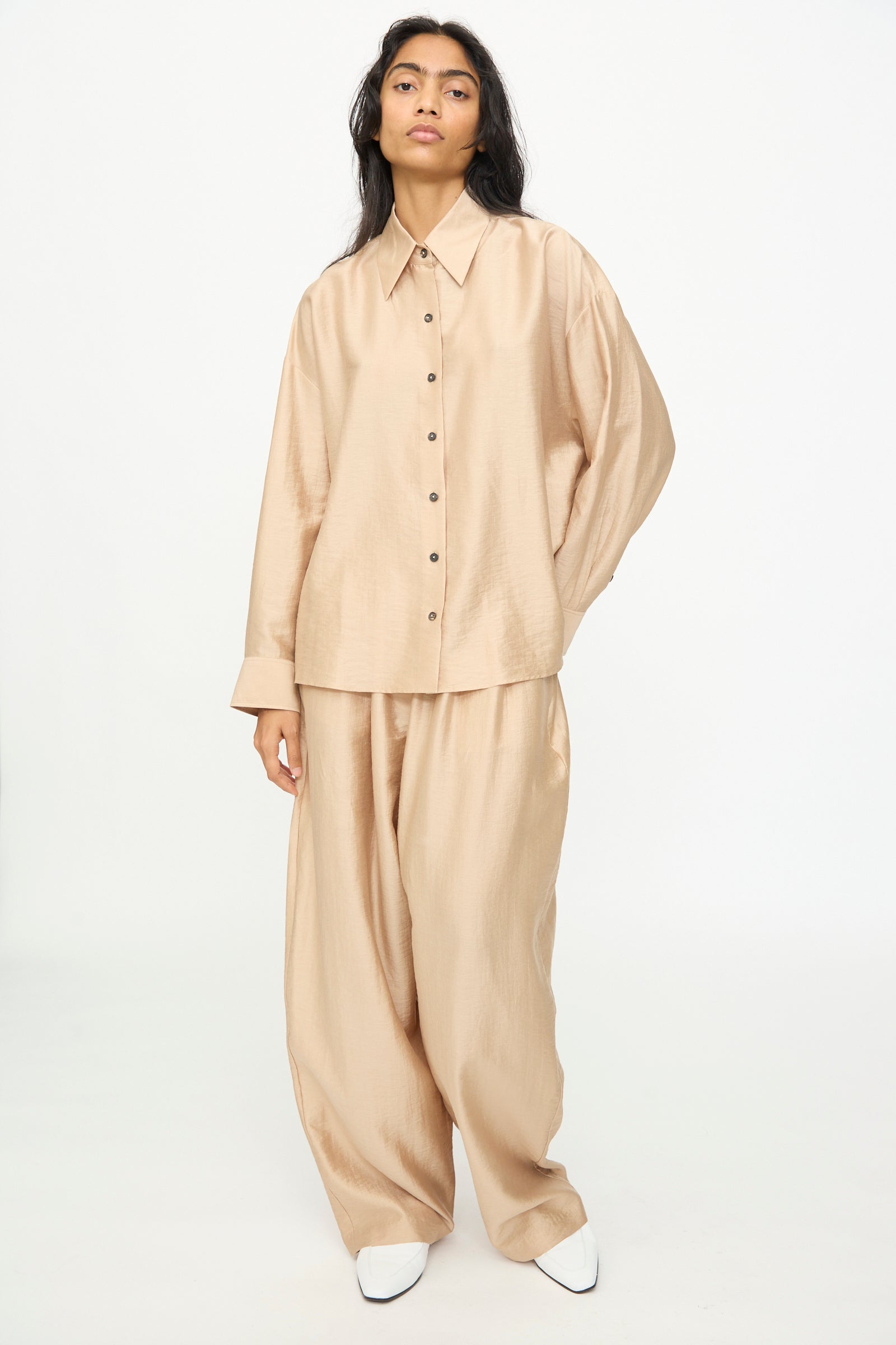 A person wearing Cordera’s Oversized Shirt in Beige, featuring a relaxed Italian collar and paired with matching wide-leg pants made from a soft lyocell blend, stands against a plain white background with white pointed shoes visible.