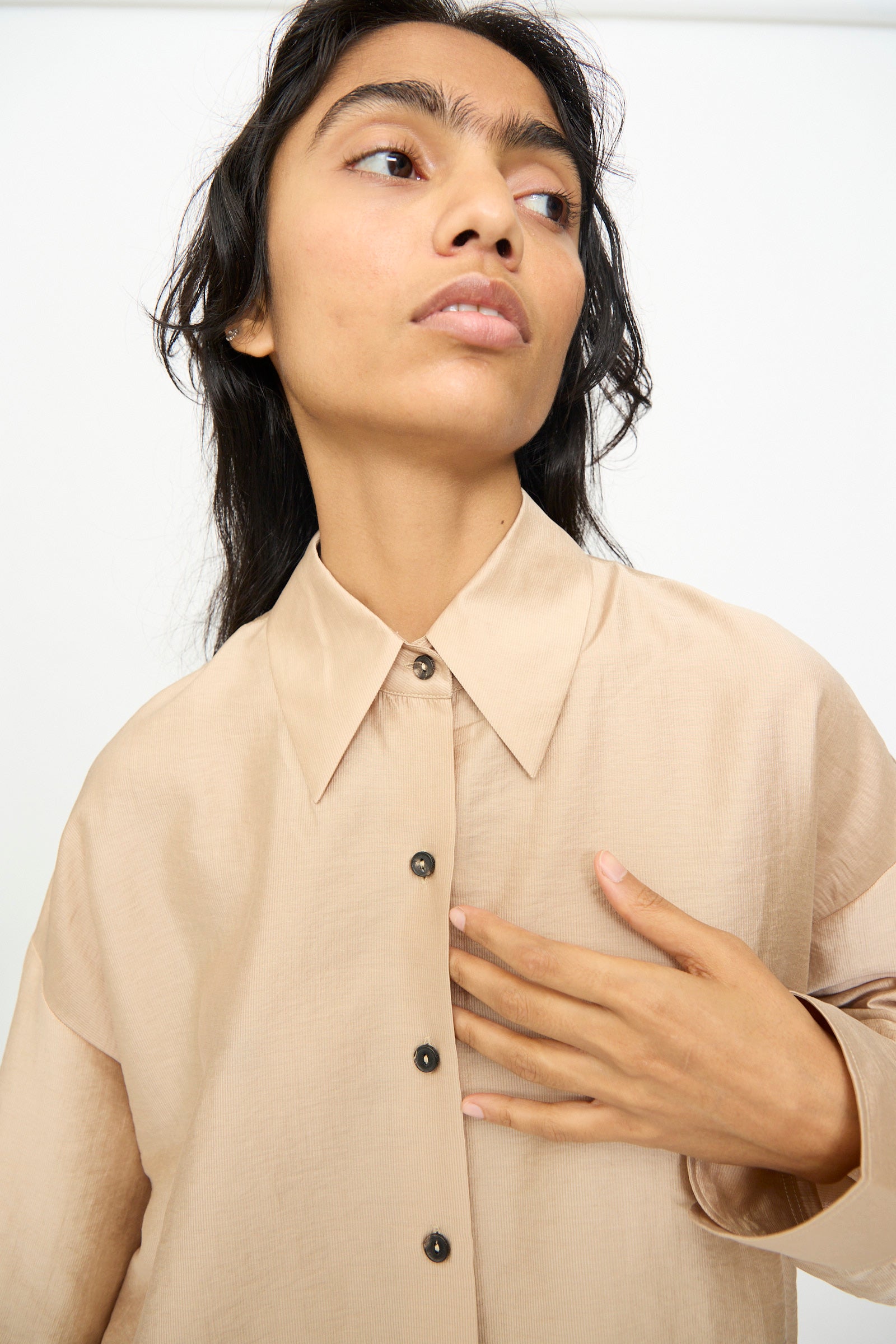 A person wears the Cordera Oversized Shirt in Beige, featuring an Italian collar and button-up design, gazing slightly upward and touching their chest with one hand against a plain background.