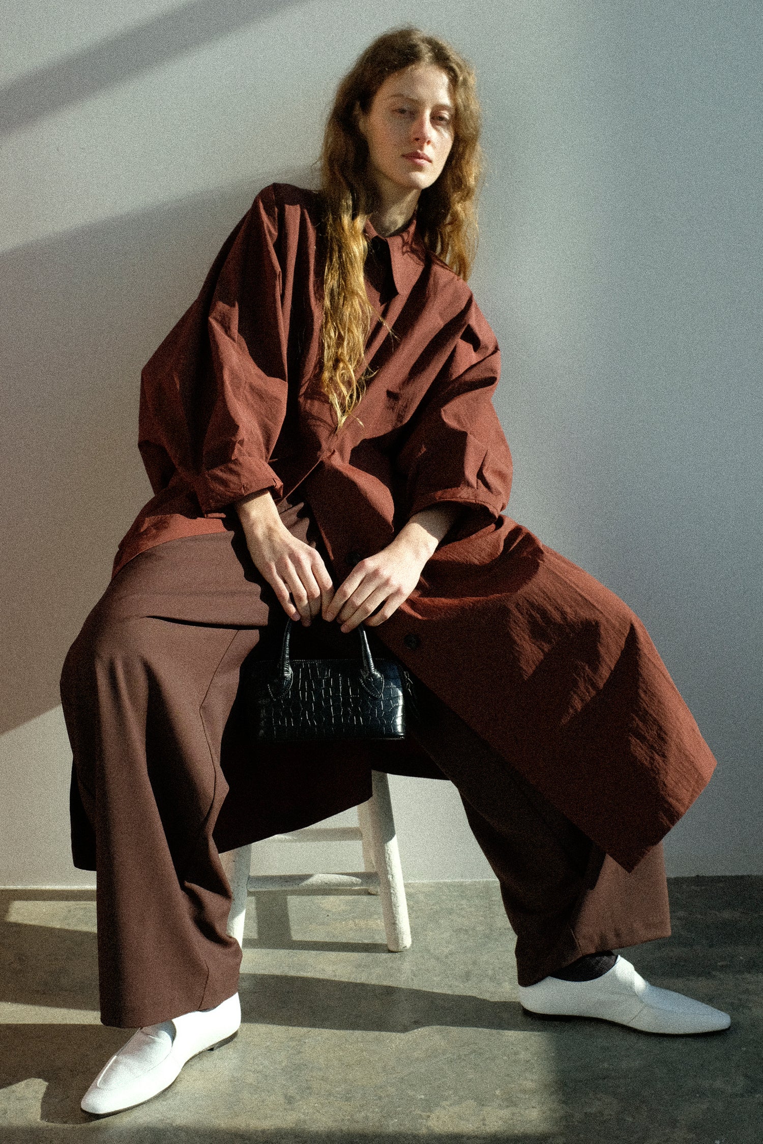 A woman wearing Cordera's Batwing Trench in Sienna, with the Tailored Crossover Pant in Burgundy, and holding the Mini Bag Crocodile in Black, sitting on a stool.