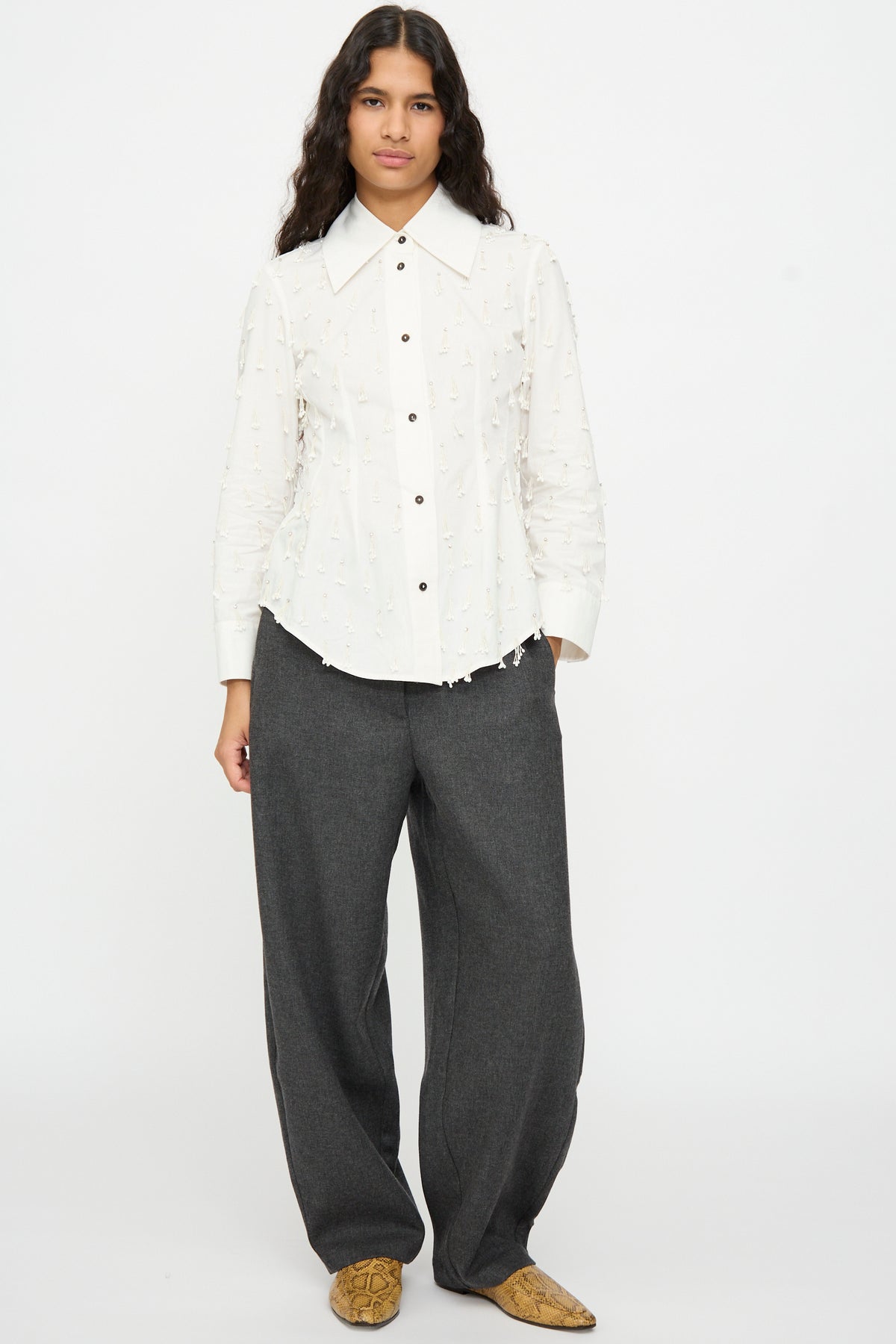 A person wearing a Cordera Petalos Shirt in White with frayed fringe details, charcoal gray wide-leg trousers, and snakeskin-print pointed-toe shoes.