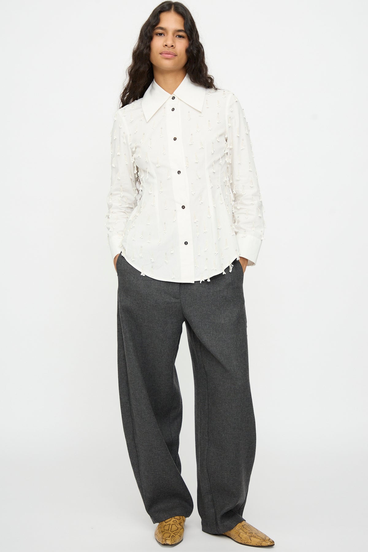 A person wearing a white Petalos Shirt with frayed accents, charcoal grey trousers with a tailored waist, and snakeskin-print loafers.