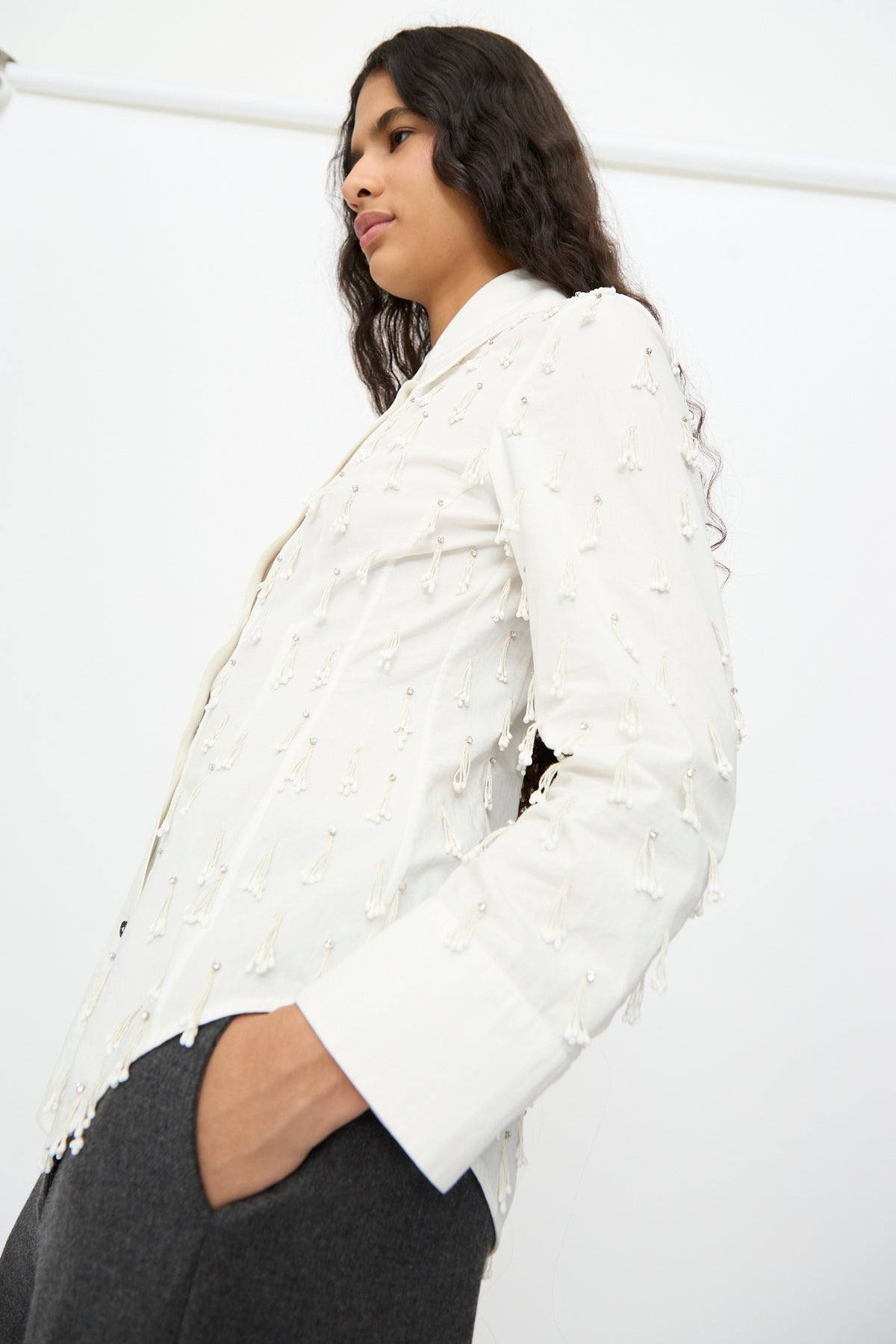 A model wearing a Cordera Petalos Shirt in White, a fitted long-sleeved top with a tailored waist and dangling bead accents.