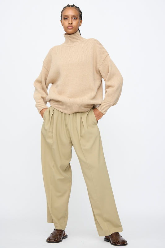 A person wearing a beige turtleneck sweater, Cordera Pleated Relaxed Pants in Olive Grey, and brown shoes stands against a plain white background.