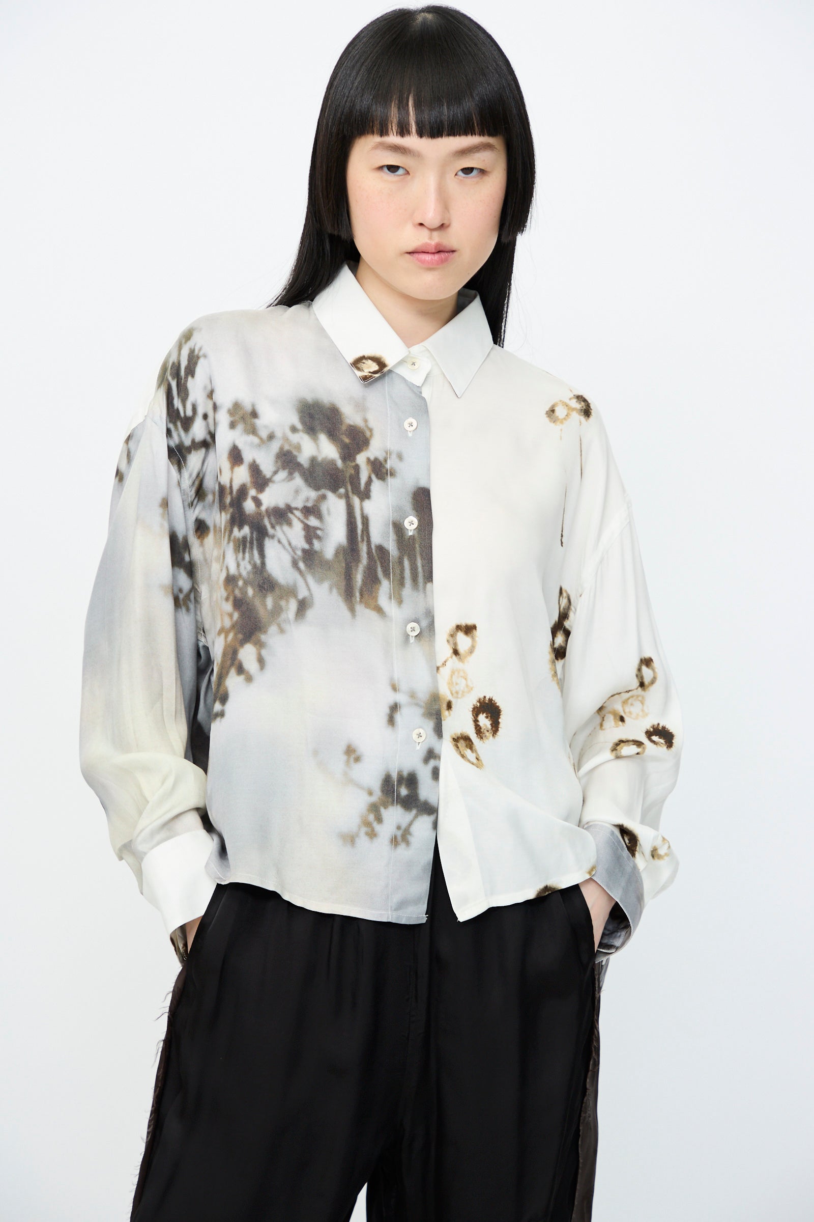 A person with straight black hair wearing an oversized Cordera Printed Classic Shirt in Bicolor, paired with black pants, stands with hands in pockets against a plain background.